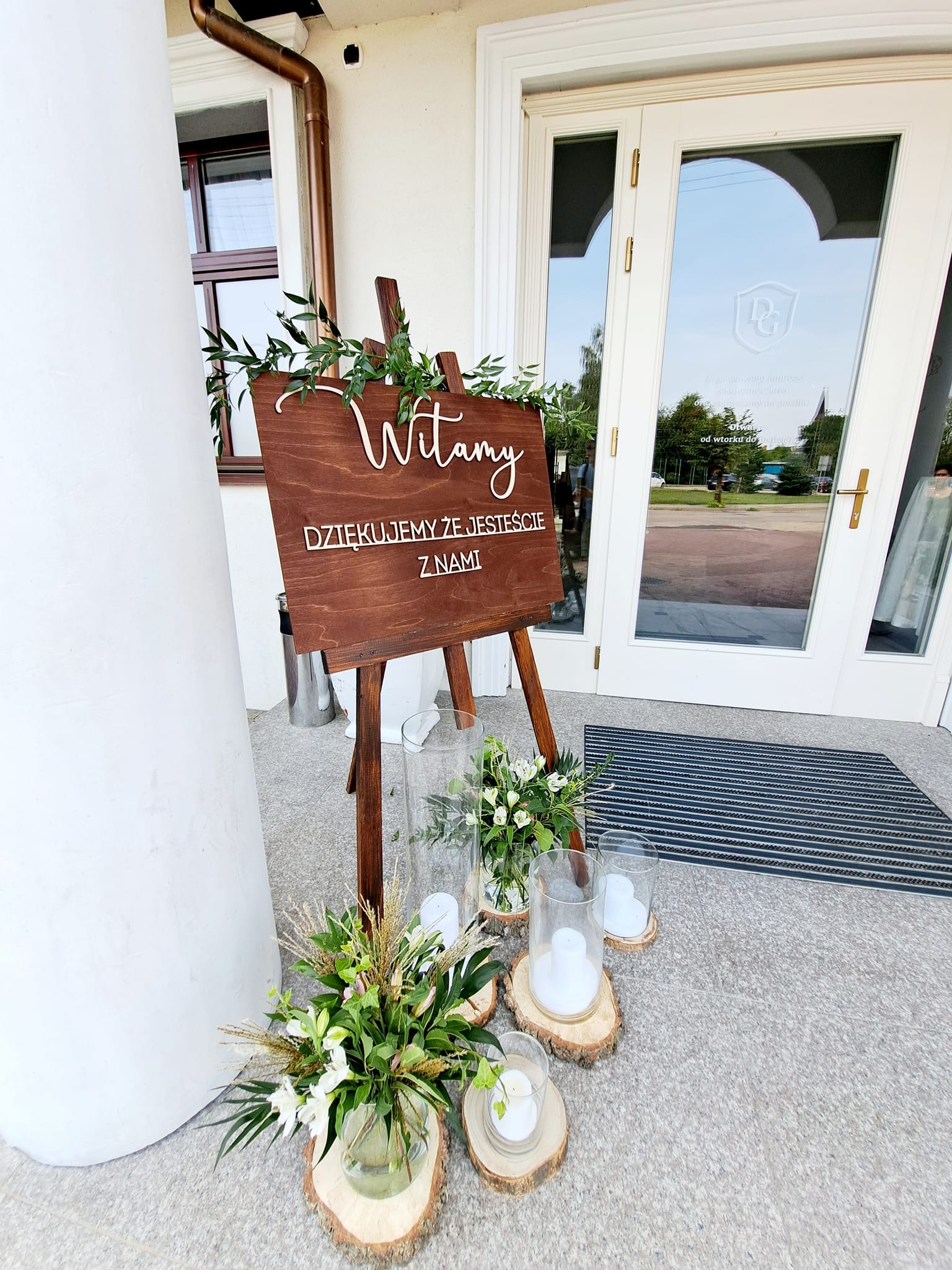 Drewniana tablica powitalna z napisem 'Witamy, dziękujemy że jesteście z nami' udekorowana zielonymi gałązkami, ustawiona przed wejściem do budynku, obok kompozycje kwiatowe i świece na drewnianych...