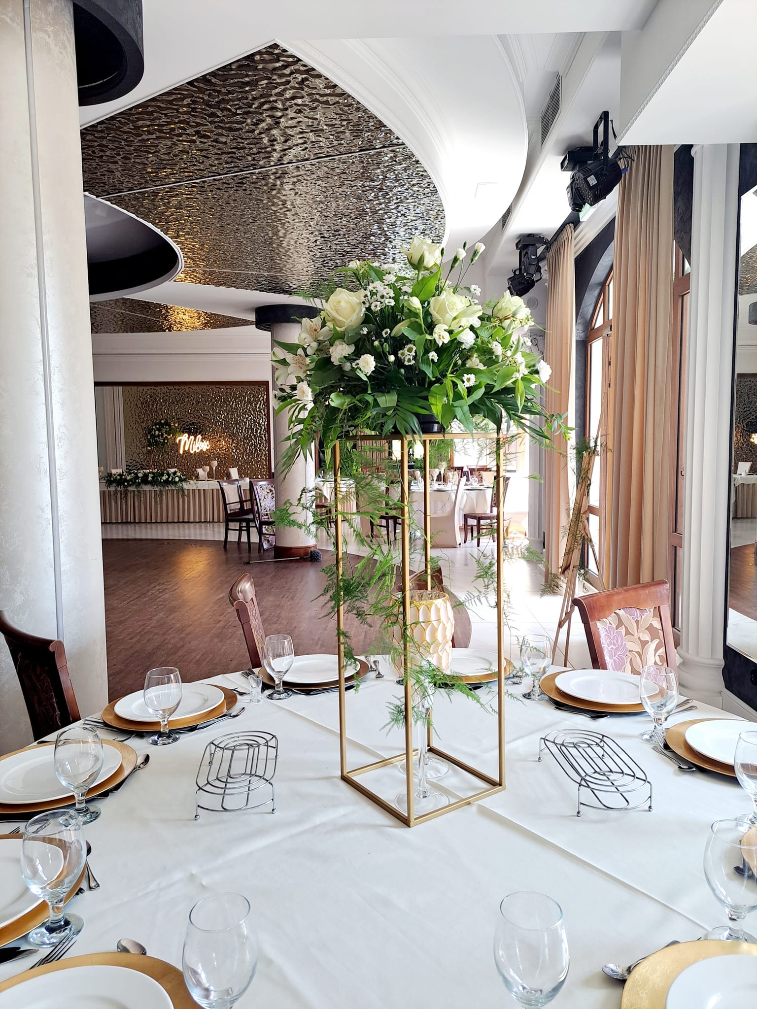 Elegancko nakryty stół z białym obrusem, złotymi podkładkami pod talerze i wysoką kompozycją kwiatową w złotej, geometrycznej konstrukcji. W tle widoczne oświetlenie sceniczne i neonowy napis...