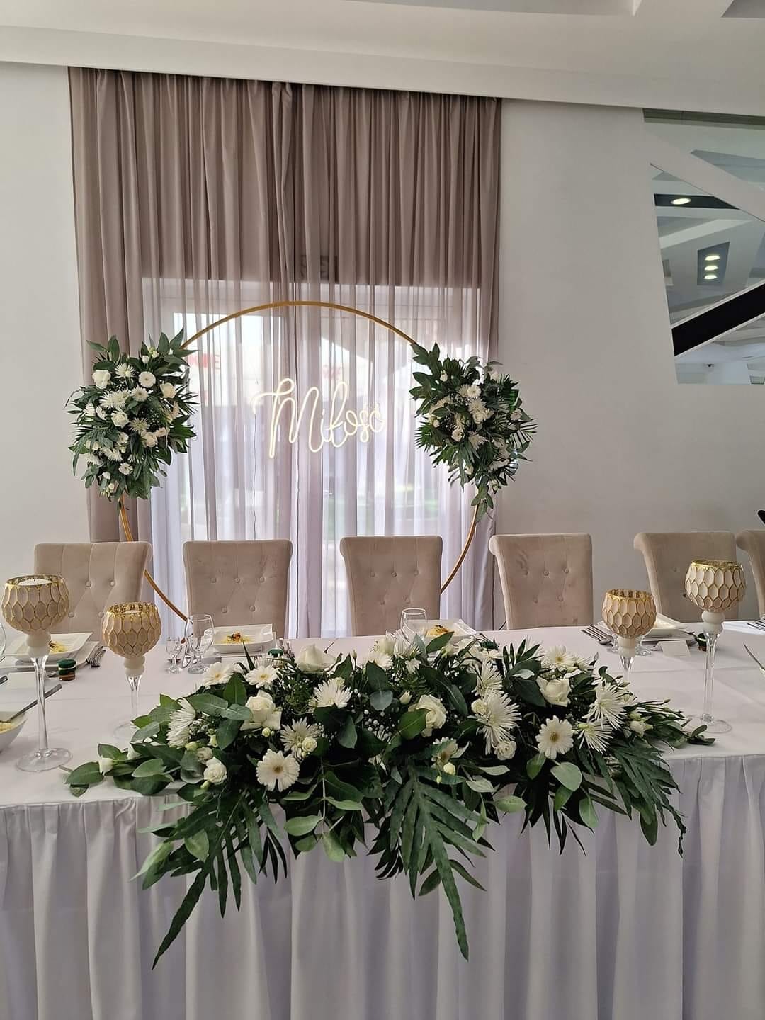 Uroczysty stół weselny z białym obrusem, kwiatową kompozycją z białych kwiatów i zielonych liści, złotym okręgiem z napisem i beżowymi krzesłami w tle.