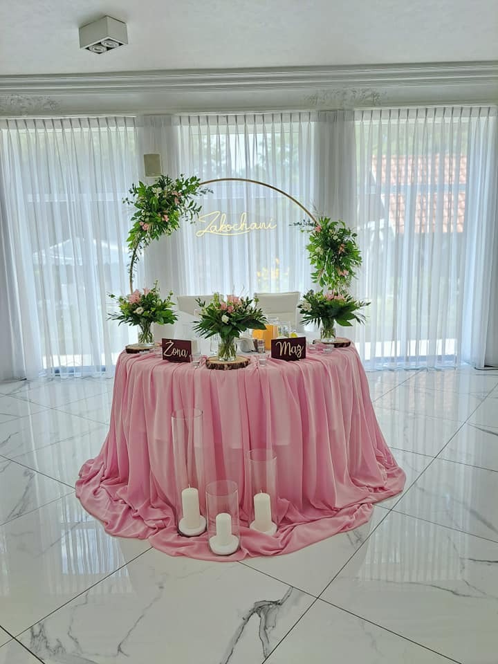 Dekoracja stołu weselnego z różowym obrusem, złotym łukiem z napisem 'Zakochani', kwiatami w drewnianych podstawkach, świecami w szklanych cylindrach oraz tabliczkami 'Żona' i 'Mąż'.