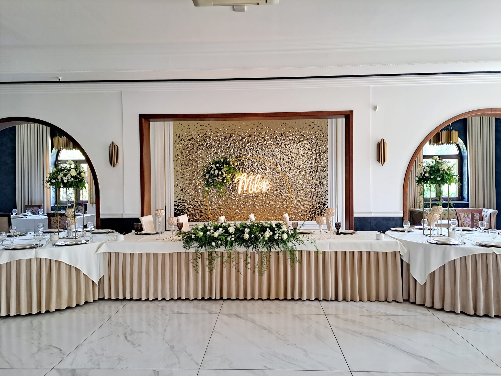 Uroczysta dekoracja stołu weselnego z kwiatami, świecami i złotym tłem z napisem 'Miłość' w eleganckiej sali z marmurową podłogą.