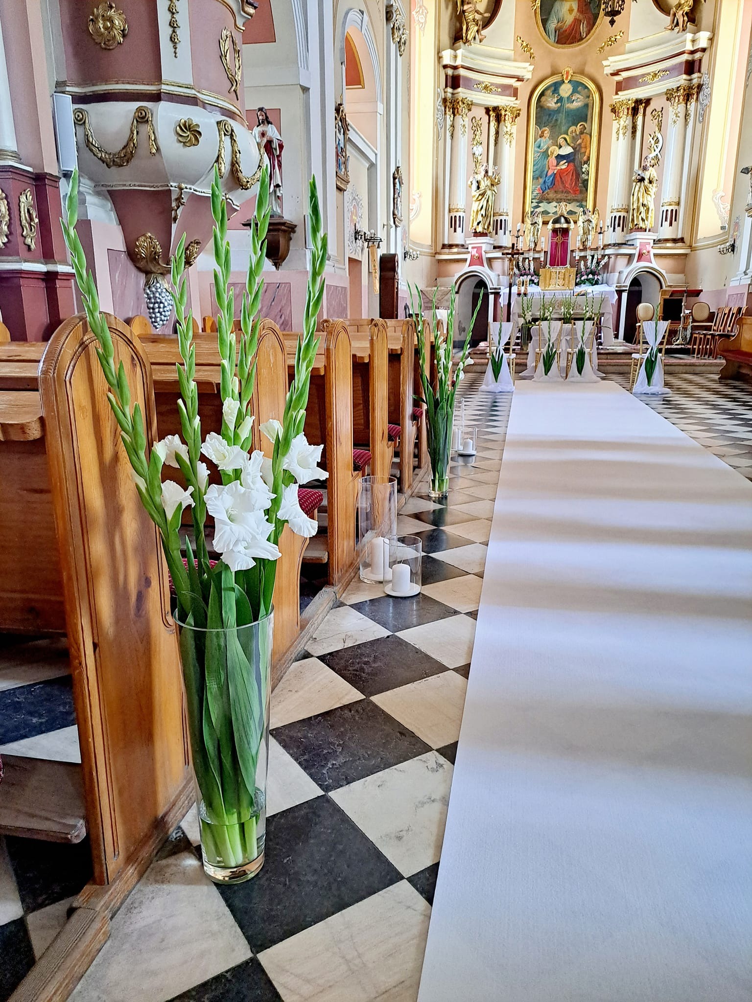 Dekoracja kwiatowa z białych mieczyków w szklanym wazonie, ustawiona w kościele obok drewnianej ławki i białego dywanu prowadzącego do ołtarza.
