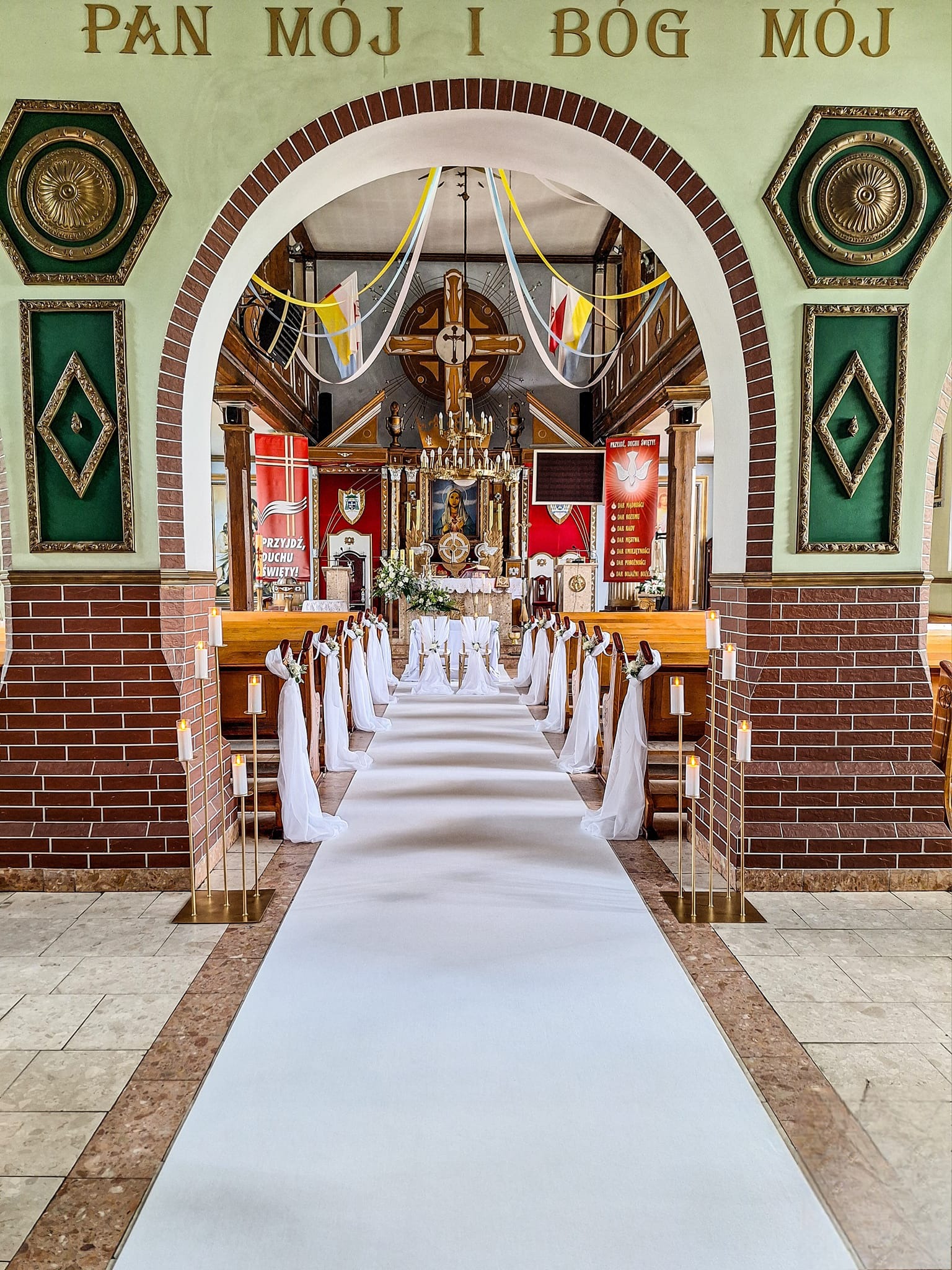 Dekoracja kościoła przed uroczystością ślubną: biały dywan, świece na złotych stojakach, girlandy z białego materiału na ławkach, ołtarz w tle.