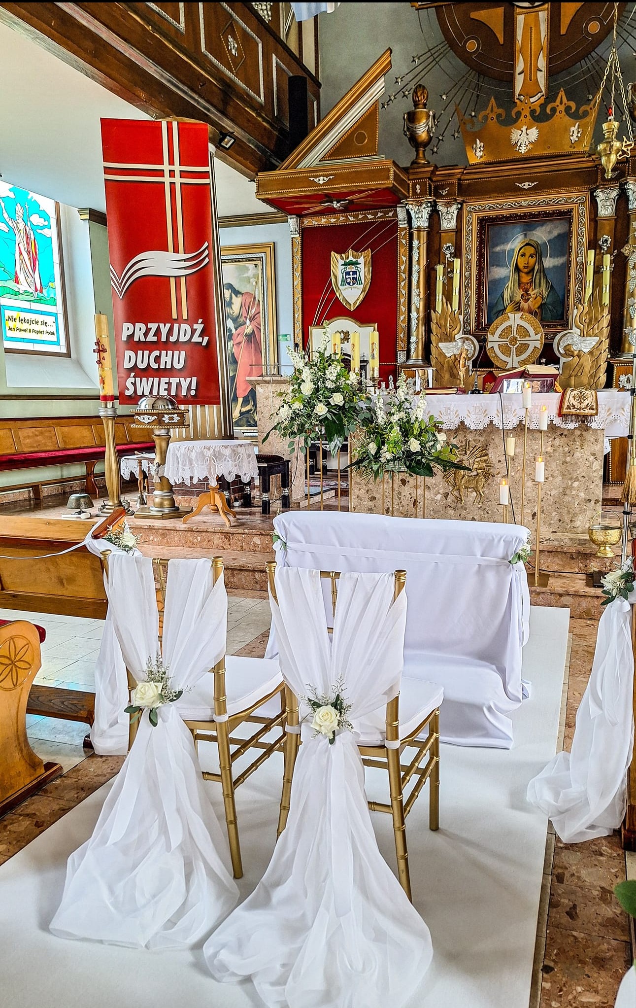 Dekoracja kościoła na uroczystość ślubną: złote krzesła z białymi draperiami i kwiatami, ołtarz z kwiatową kompozycją, czerwony baner z napisem 'Przyjdź Duchu Święty!'