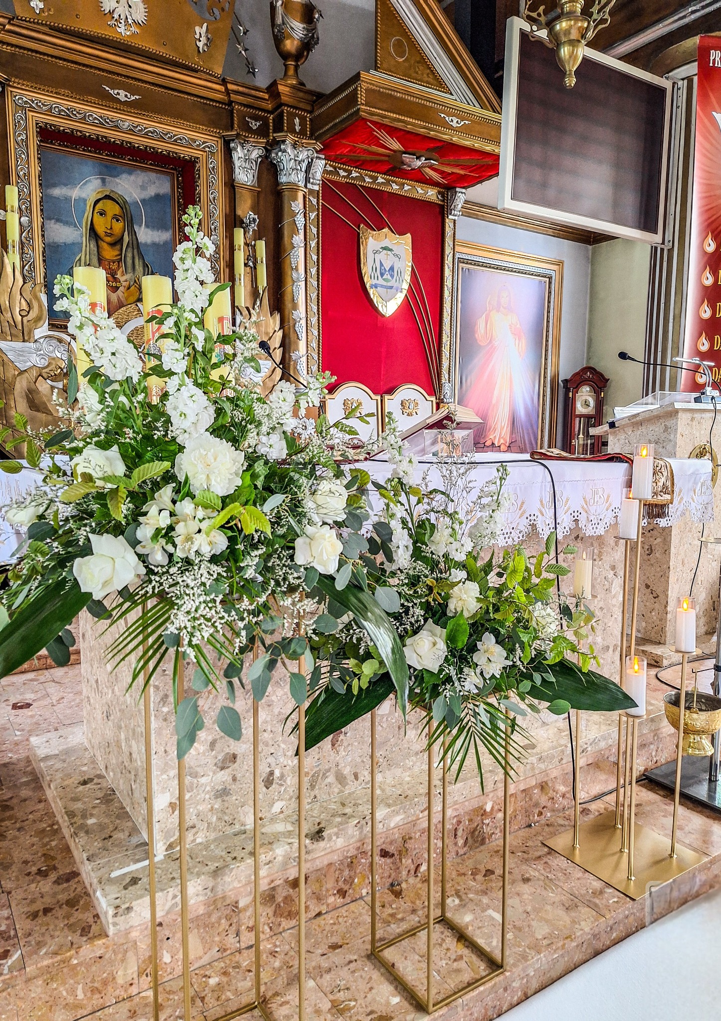 Dekoracja kwiatowa z białych róż i zielonych liści na złotych stojakach w kościele, z ołtarzem i obrazami w tle.