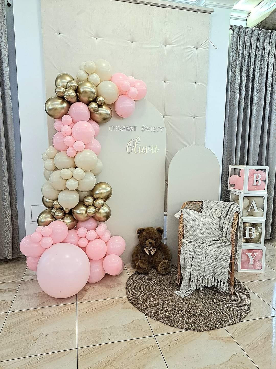 Dekoracja na Chrzest Święty Oliwii: girlanda z różowych, złotych i kremowych balonów, pluszowy miś, wiklinowy fotel z pledem, białe pudełka z napisem BABY.
