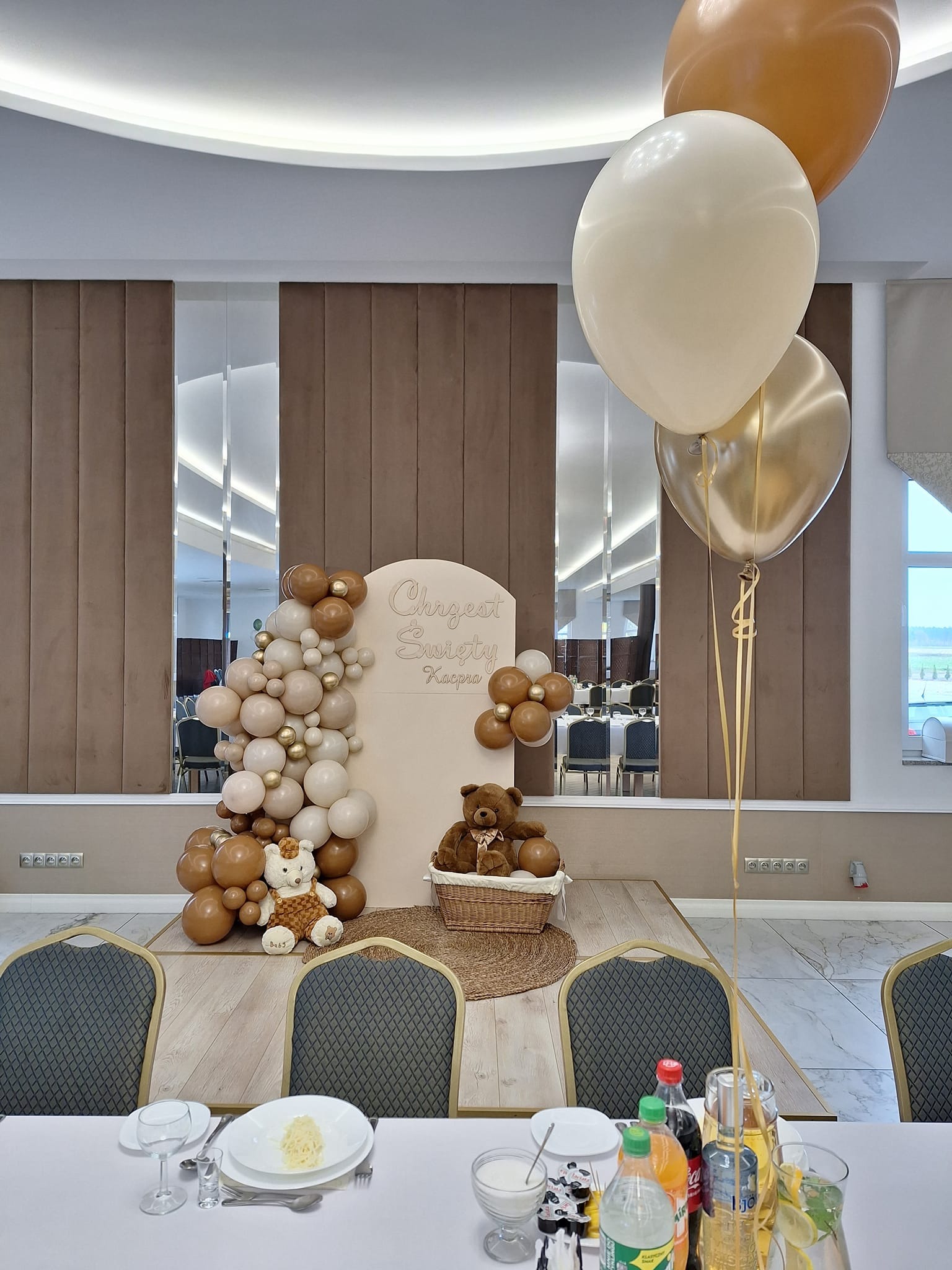 Dekoracja na Chrzest Święty: beżowe i złote balony, pluszowe misie, ścianka z napisem 'Chrzest Święty Kacpra' w sali bankietowej, nakryty stół z napojami i talerzami.