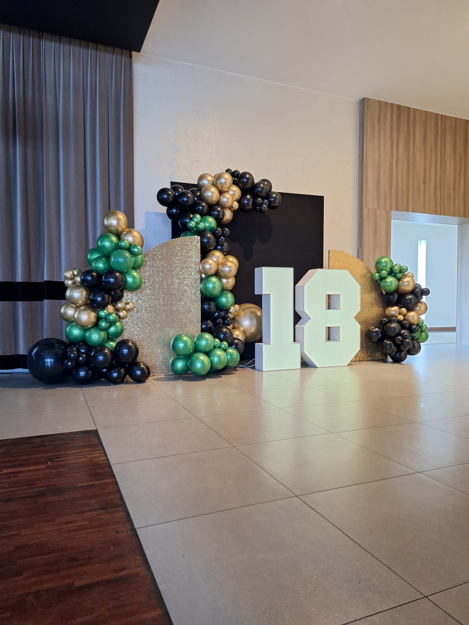 Dekoracja urodzinowa z białymi cyframi 18, balonami w kolorach czarnym, złotym i zielonym oraz błyszczącymi panelami na tle jasnego wnętrza.