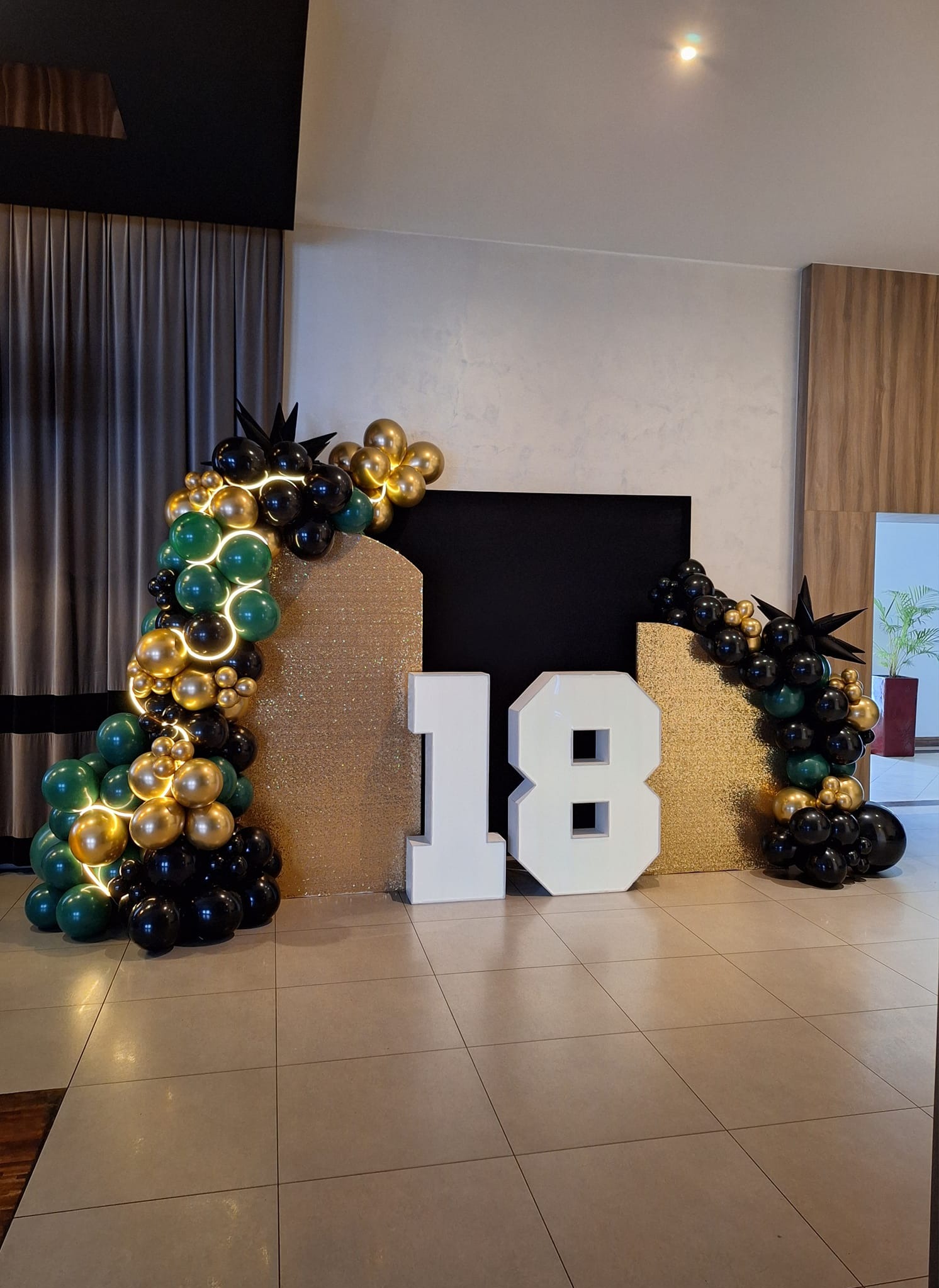 Dekoracja na 18 urodziny: białe cyfry '18' otoczone łukami z czarnych, złotych i zielonych balonów z delikatnym podświetleniem, na tle złotych ścianek i czarnego panelu.