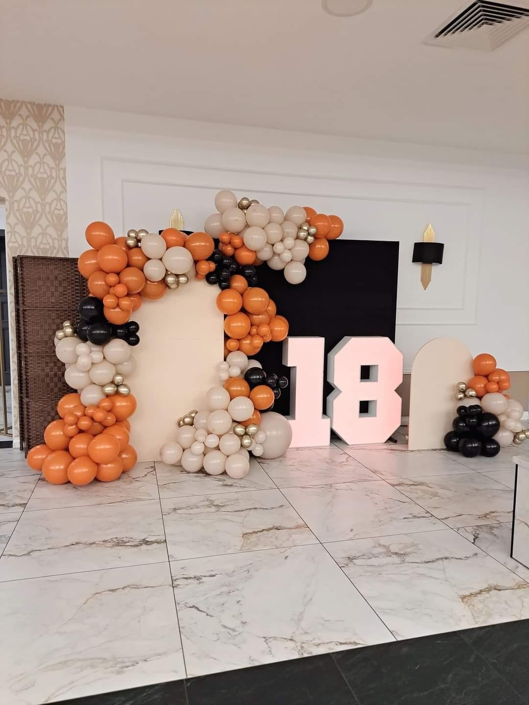 Dekoracja urodzinowa z balonów w kolorach pomarańczowym, białym i czarnym, ułożonych w kształt łuku wokół białych paneli i cyfr '18' podświetlonych na różowo.