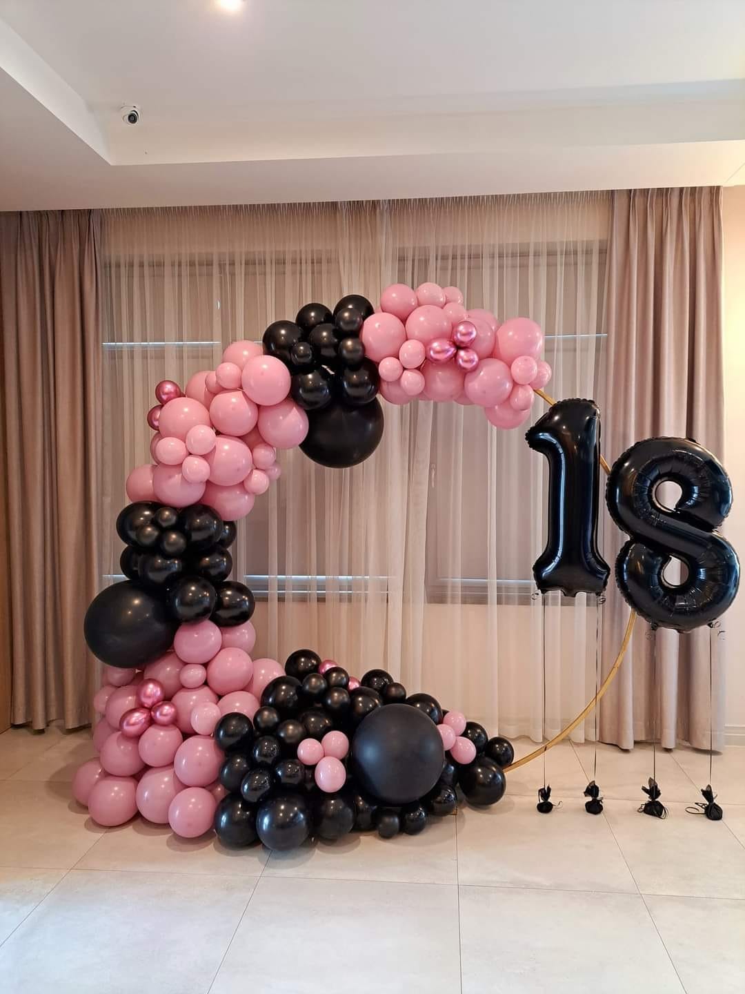 Dekoracja balonowa na osiemnaste urodziny: łuk z różowych i czarnych balonów oraz cyfry '18' z czarnych balonów foliowych, całość na tle beżowych zasłon.