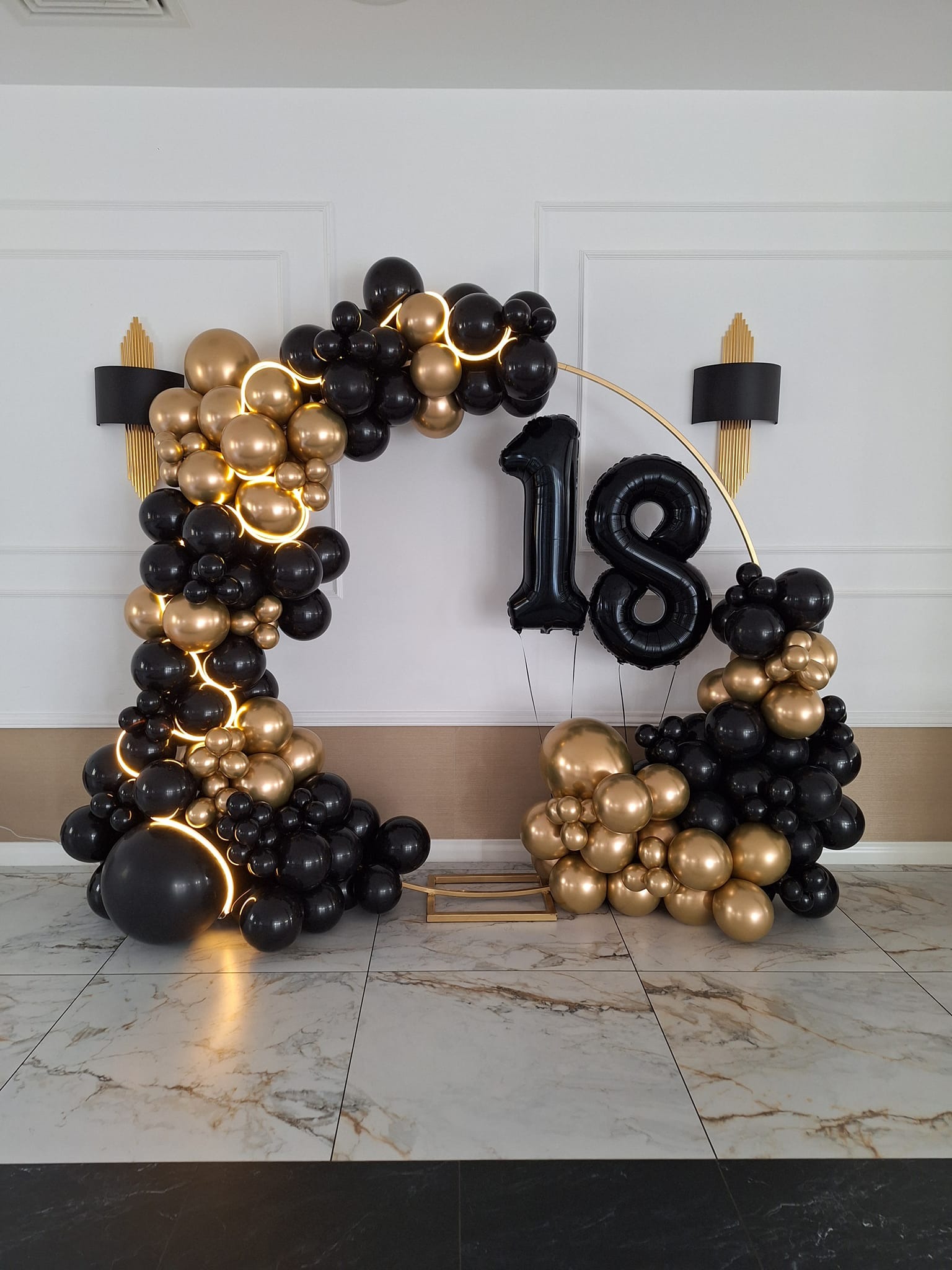 Dekoracja balonowa na 18 urodziny: czarne i złote balony ułożone w łuk z podświetleniem, cyfry 1 i 8 z balonów foliowych, marmurowa podłoga.