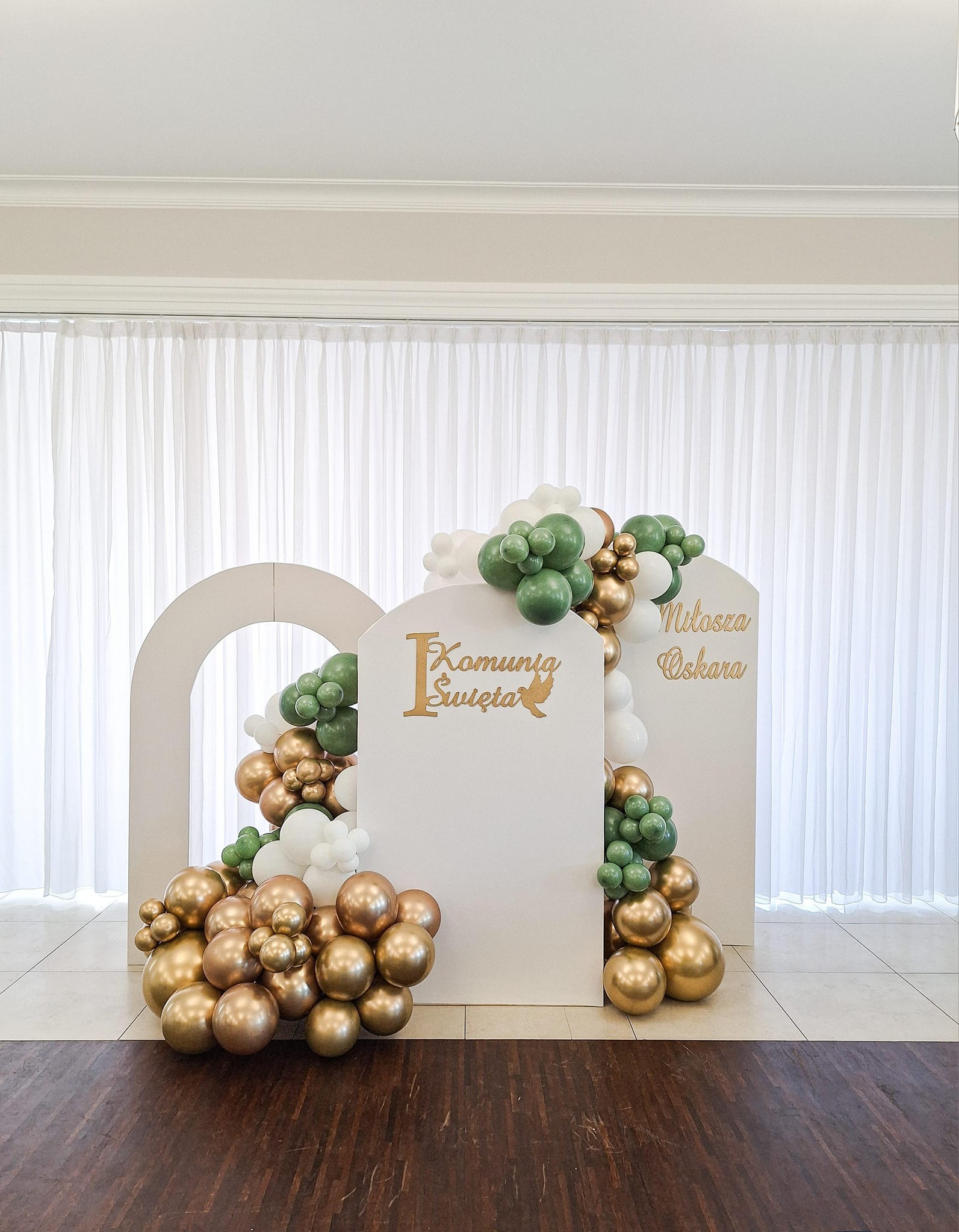 Dekoracja balonowa w odcieniach złota, zieleni i bieli, zdobiąca białe ścianki z napisem 'I Komunia Święta' oraz imiona, na tle białych zasłon.