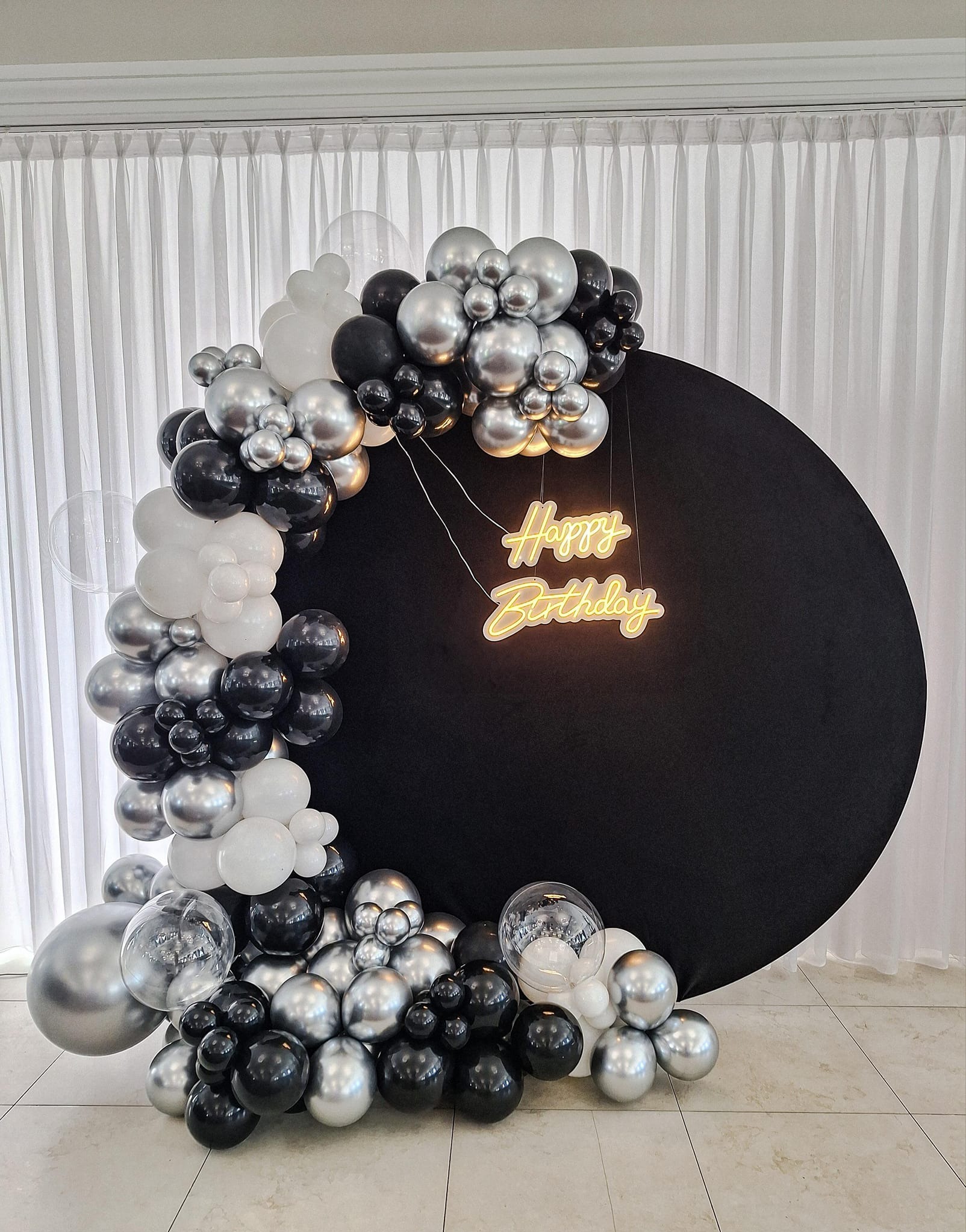 Dekoracja urodzinowa z czarnym, okrągłym tłem, neonowym napisem 'Happy Birthday' i girlandą z czarnych, srebrnych i białych balonów.