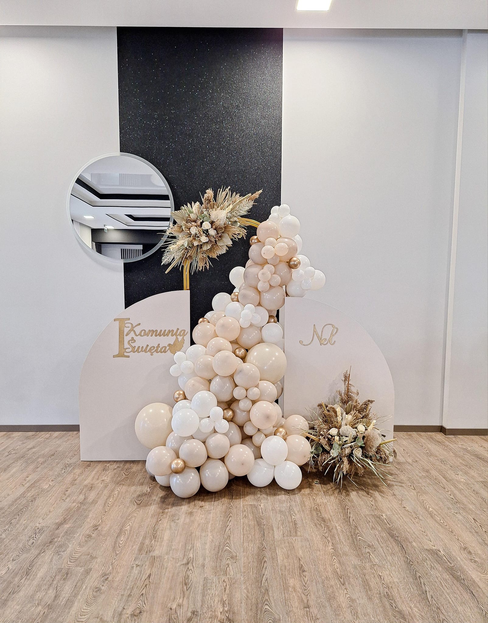 Dekoracja balonowa w odcieniach bieli i beżu z elementami złota, uzupełniona suchymi kwiatami na tle paneli z napisem 'Komunia Święta' i lustrem.