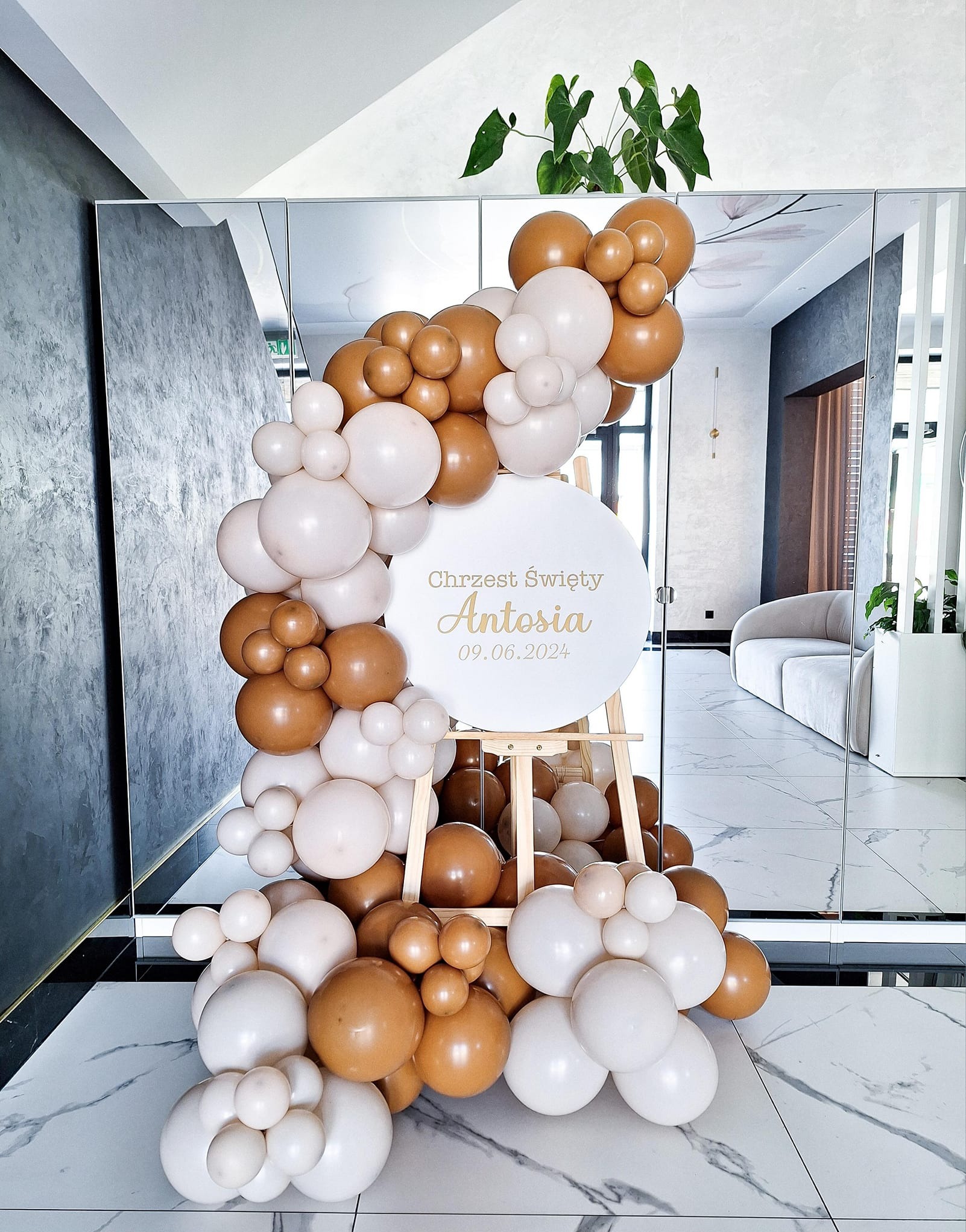 Dekoracja balonowa w odcieniach brązu i bieli, z napisem 'Chrzest Święty Antosia 09.06.2024' na okrągłej tablicy umieszczonej na drewnianej sztaludze, ustawiona w eleganckim wnętrzu w Brzezinach.