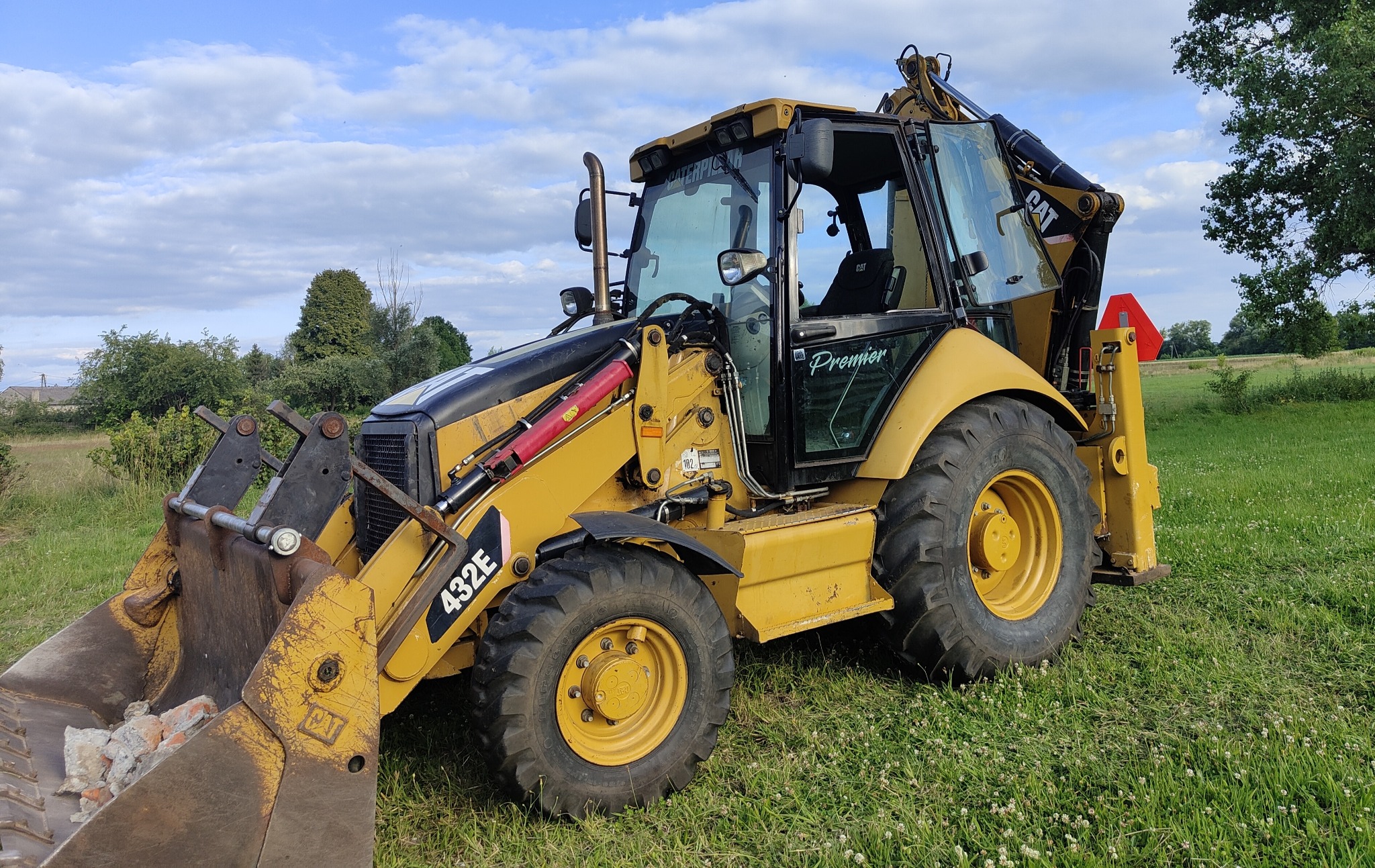 Żółta koparko-ładowarka Caterpillar 432E z łyżką wypełnioną gruzem na zewnątrz, widoczny trójkąt ostrzegawczy na tylnej części maszyny.