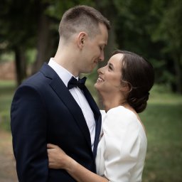 Iza Wiklina - Uśmiechnięta panna młoda w białej sukni obejmuje pana młodego w granatowym smokingu, patrząc sobie w oczy na tle zieleni.