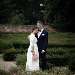 Iza Wiklina - Elegancka sesja ślubna w ogrodzie: Panna Młoda w białej sukni z bukietem, Pan Młody w granatowym smokingu, czule obejmujący ukochaną. Romantyczna sceneria parkowa.