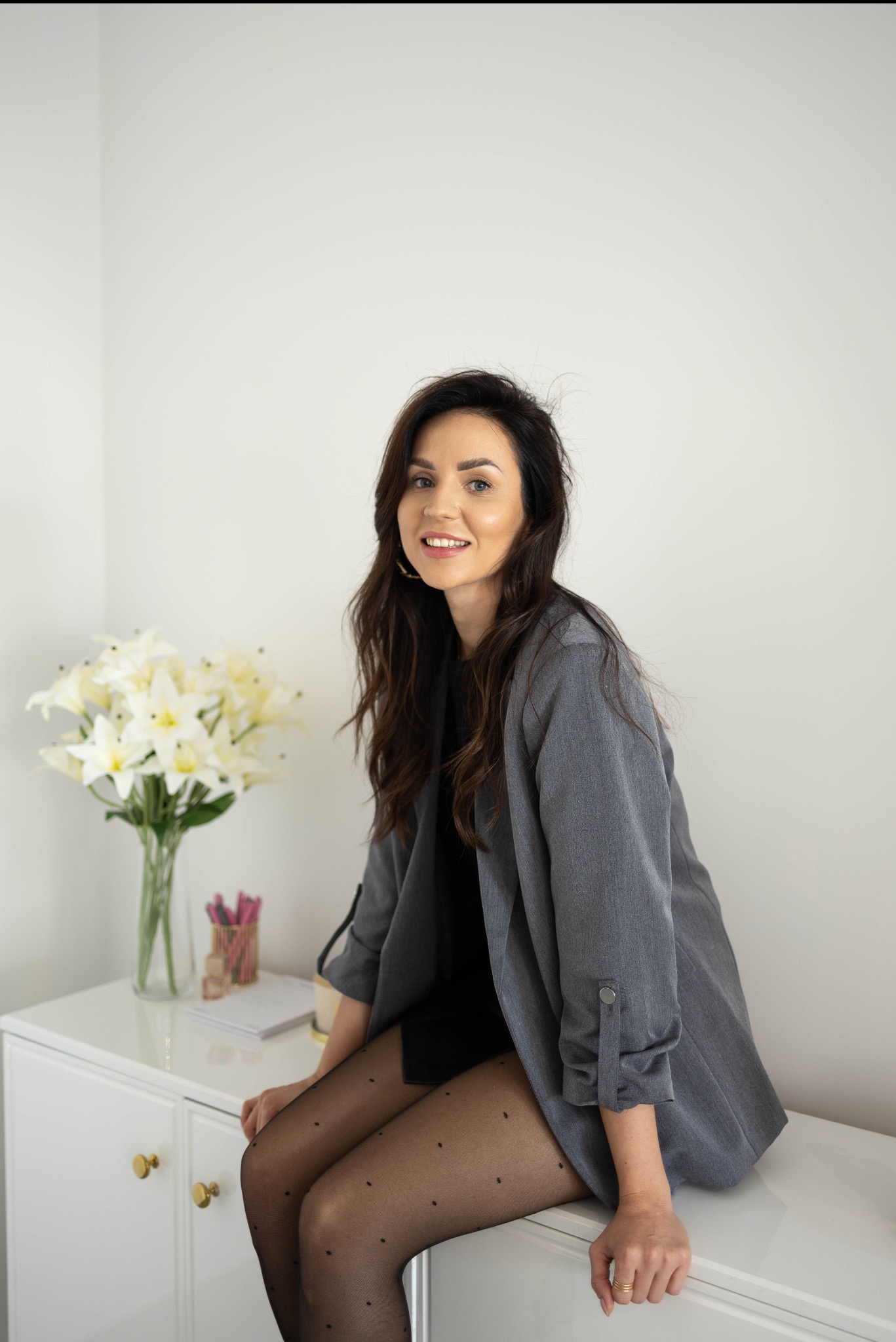 Portret uśmiechniętej kobiety w szarym żakiecie, siedzącej na białej komodzie z bukietem lilii w tle. Sesja wizerunkowa z subtelnym oświetleniem.