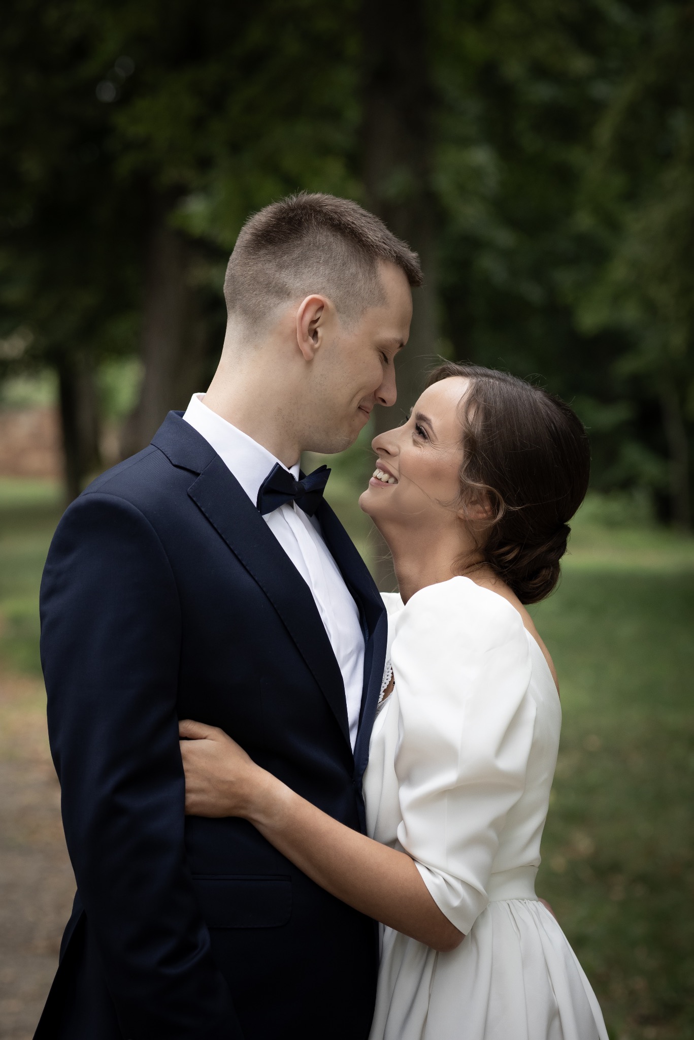 Uśmiechnięta panna młoda w białej sukni obejmuje pana młodego w granatowym smokingu, patrząc sobie w oczy na tle zieleni.