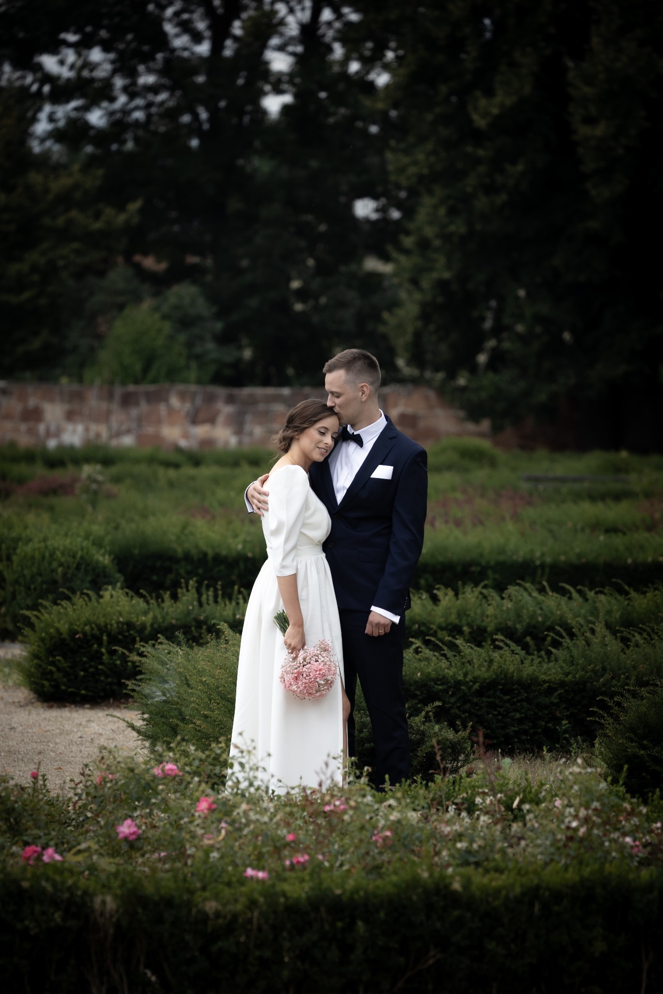 Elegancka sesja ślubna w ogrodzie: Panna Młoda w białej sukni z bukietem, Pan Młody w granatowym smokingu, czule obejmujący ukochaną. Romantyczna sceneria parkowa.
