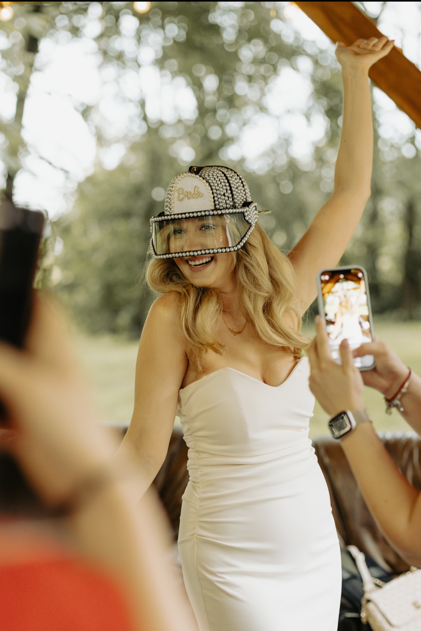 Uśmiechnięta blondynka w białej sukience i czapce z napisem 'Bride', otoczona osobami robiącymi zdjęcia telefonami, na tle zieleni. Ujęcie z wieczoru panieńskiego.