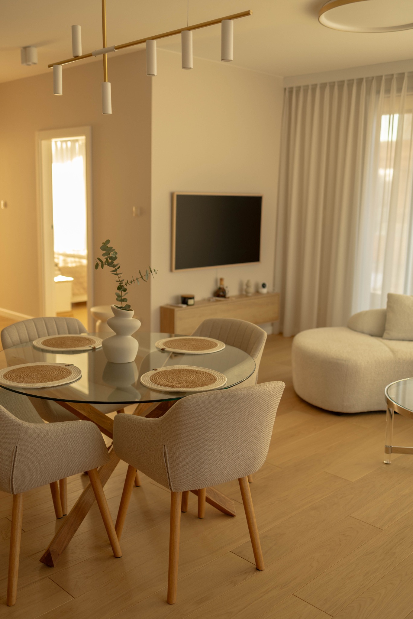 Jasne wnętrze apartamentu: okrągły stół z blatem ze szkła, beżowe krzesła, dekoracje. W tle telewizor i sofa. Stonowana kolorystyka, naturalne światło.