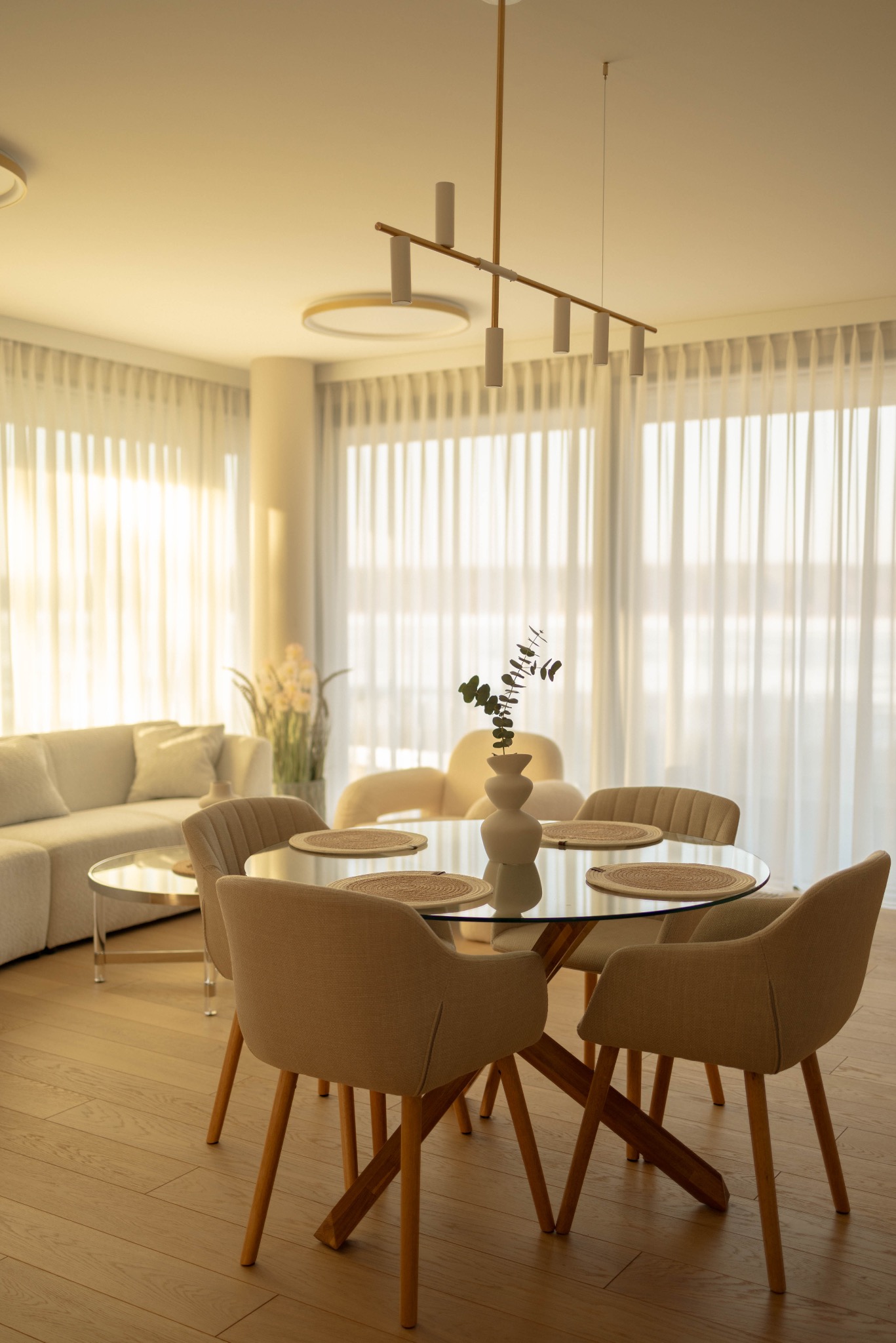 Jasne wnętrze apartamentu: okrągły stół z szklanym blatem, beżowe krzesła, sofa przy oknie z białymi zasłonami. Uwagę przyciąga minimalistyczna lampa sufitowa.