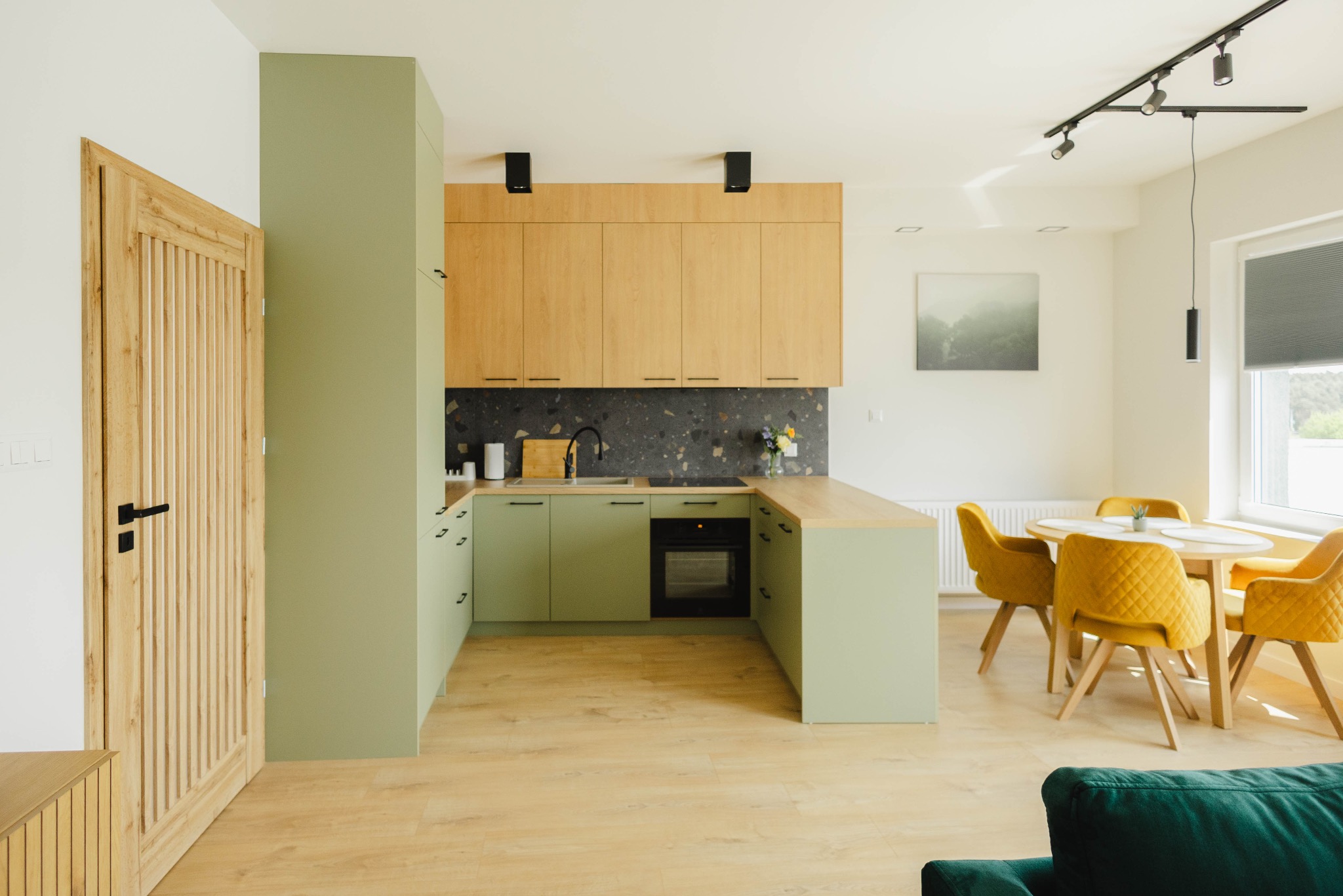 Jasne, nowoczesne wnętrze: kuchnia z zielonymi szafkami, drewnianymi blatami i żółtymi krzesłami. Ujęcie szerokokątne, podkreślające przestronność mieszkania.