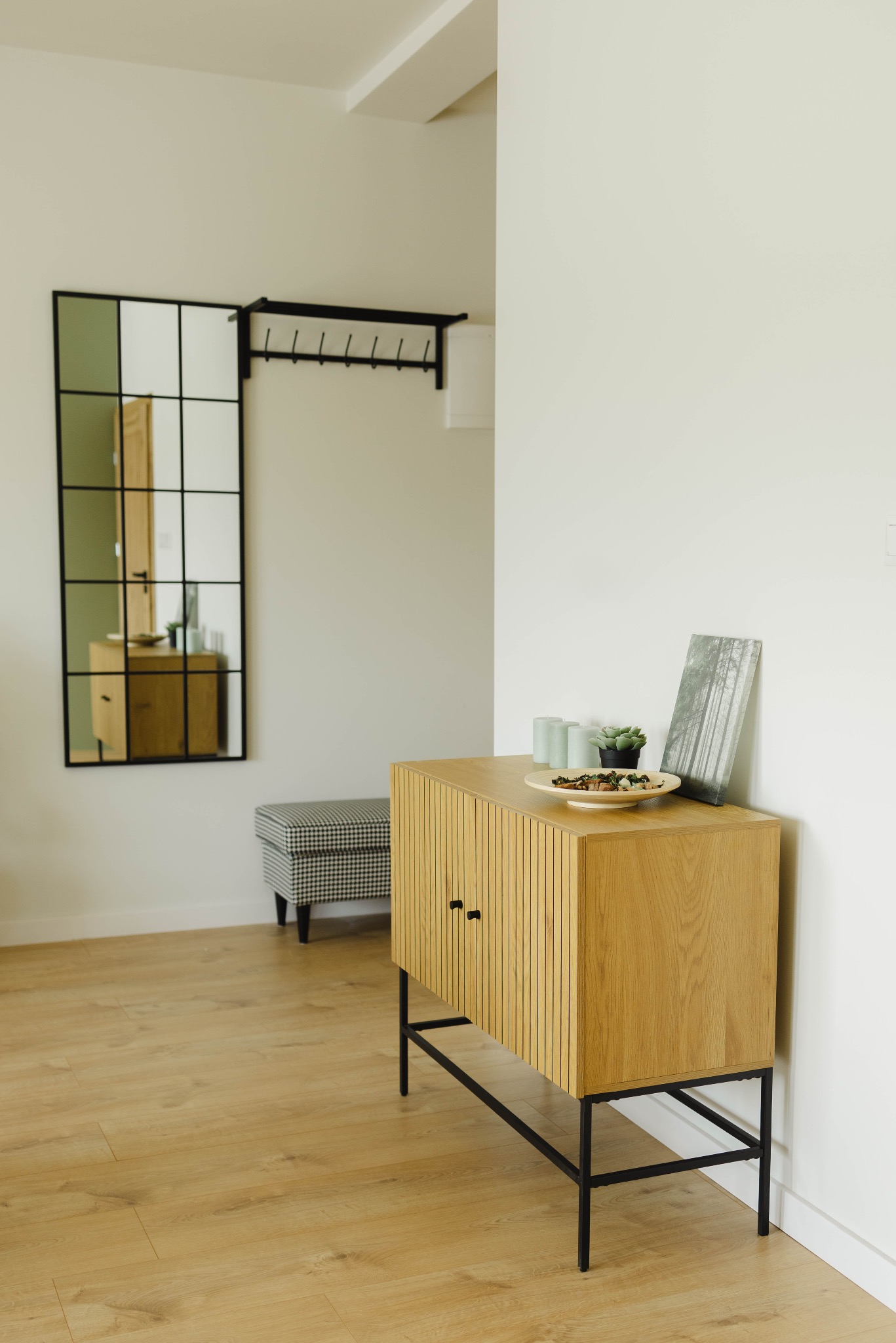 Jasne wnętrze: komoda z dekoracjami, lustro z czarną ramą i wieszak na ubrania. Podłoga drewniana, neutralne kolory ścian. Elementy dekoracyjne na komodzie.