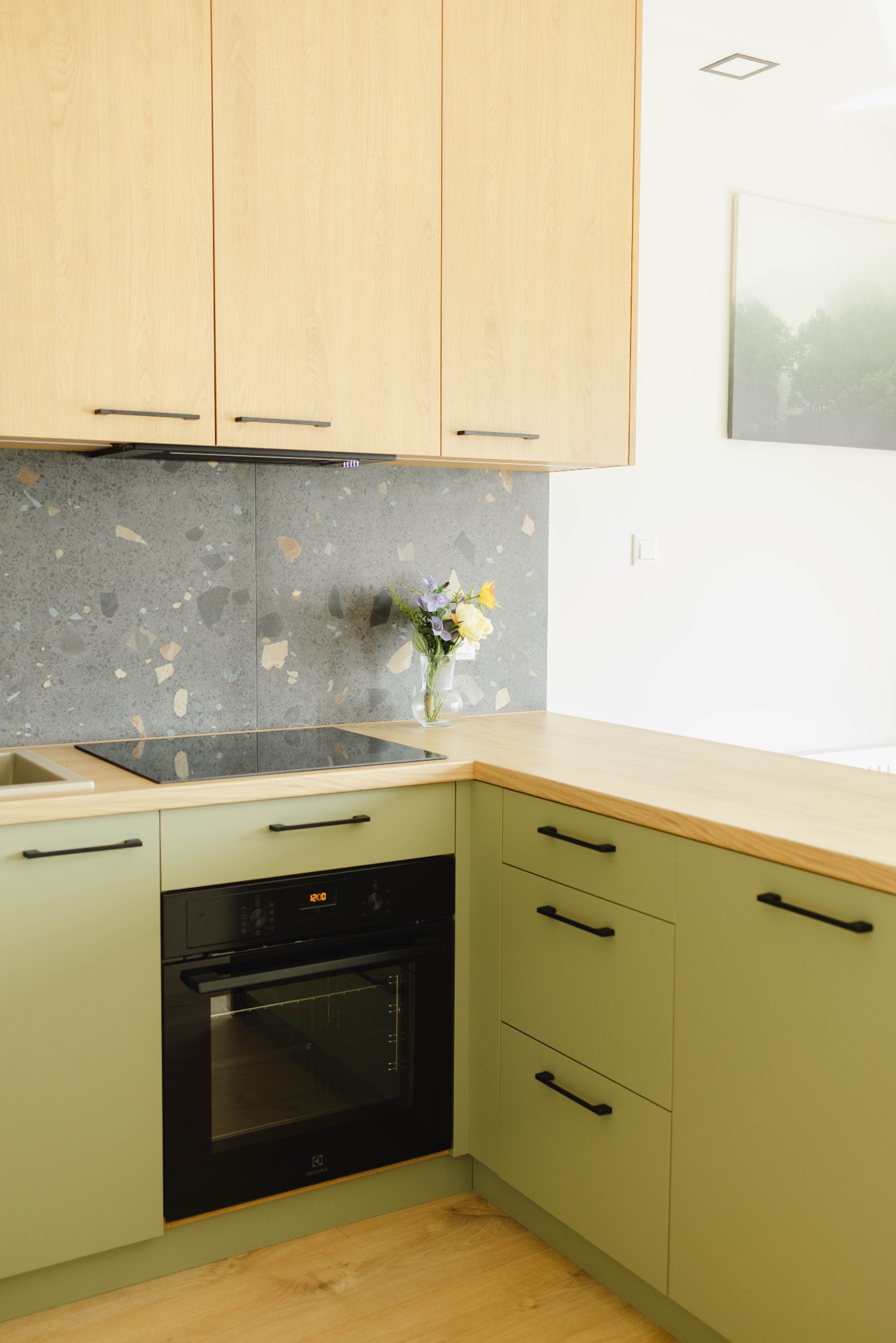 Jasna kuchnia w odcieniach zieleni i drewna z blatem, kwiatami w wazonie i płytą indukcyjną na tle ściany z mozaiką. Nowoczesny design, oświetlenie punktowe.