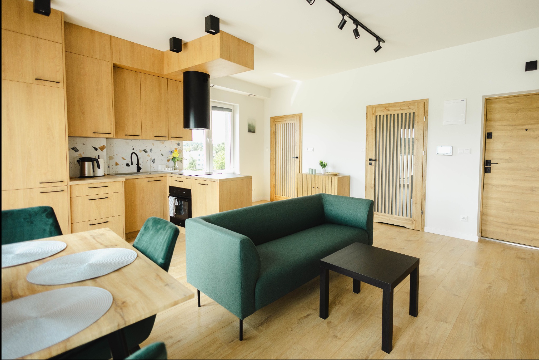 Jasne wnętrze mieszkania w Kielcach: drewniane meble kuchenne, zielona sofa, stół z krzesłami. Nowoczesny design, naturalne światło, parkiet. Minimalistyczny styl.