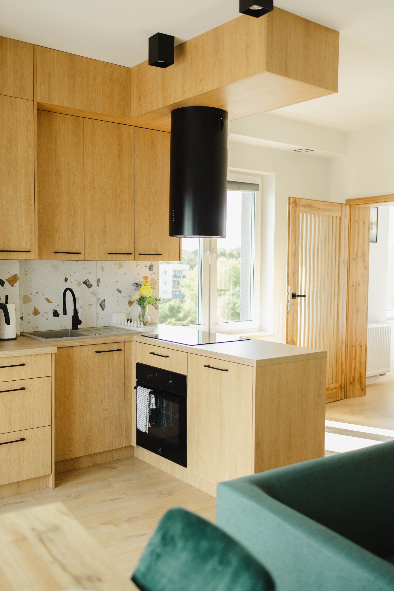 Jasna kuchnia z drewnianymi szafkami, czarnym okapem i armaturą. Na blacie wazon z kwiatami. W tle okno z widokiem na zieleń i drewniane drzwi do innego pomieszczenia.