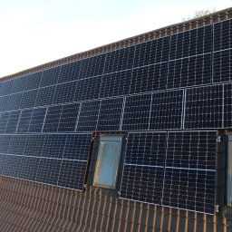 Smart Electro Cezary Mucha - Montaż paneli słonecznych na brązowym dachu z oknami dachowymi, widoczny pracownik w pomarańczowym kasku.
