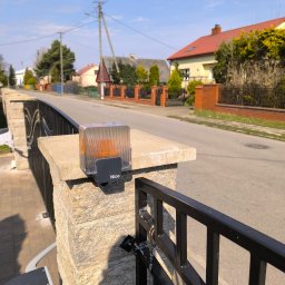 Smart Electro Cezary Mucha - Lampa ostrzegawcza Nice zamontowana na słupku bramy wjazdowej, widok na ulicę i zabudowę jednorodzinną w tle. Słoneczny dzień.