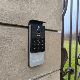 Smart Electro Cezary Mucha - Zamontowany wideodomofon STEECH z klawiaturą numeryczną na murze obok metalowej bramy wjazdowej. Widoczne zielone tło ogrodu.