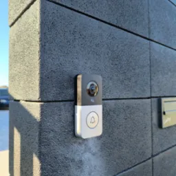 Nowoczesny wideodomofon z kamerą zamontowany na szarym, teksturowanym słupku ogrodzeniowym, z widoczną skrzynką na listy i fragmentem nieba w tle.