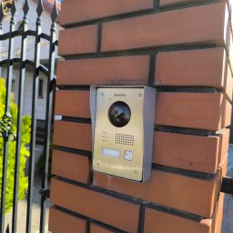Smart Electro Cezary Mucha - Złoto-srebrny wideodomofon Hikvision zamontowany na ceglanym słupku ogrodzenia, widok z boku, brama w tle, słoneczny dzień.
