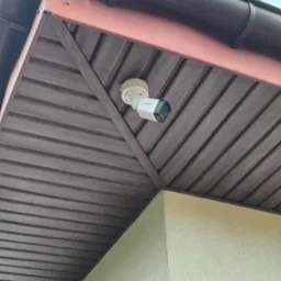 Biała kamera monitoringu Hikvision zamontowana pod brązowym podbitką dachu na zewnątrz budynku, widok z dołu.