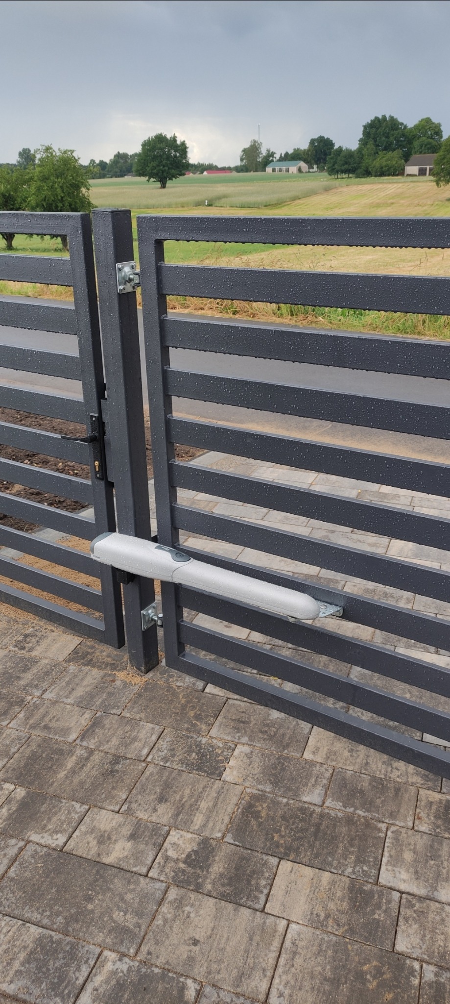 Szara, metalowa brama posesyjna z zamontowanym napędem automatycznym, widoczne krople deszczu na konstrukcji, w tle pole i zabudowania.