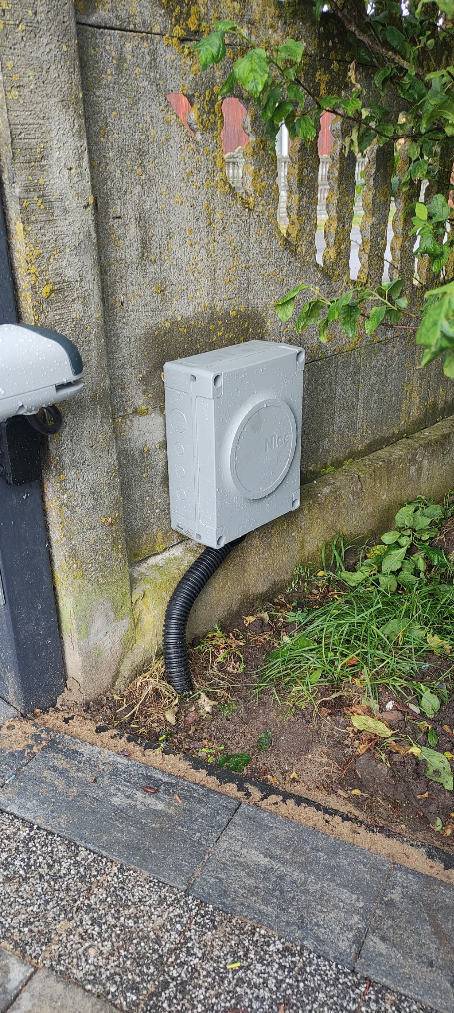 Szara skrzynka sterownicza z logo Nice, zamontowana na betonowym słupku ogrodzenia, połączona czarnym, karbowanym przewodem z urządzeniem po lewej stronie, prawdopodobnie napędem bramy.