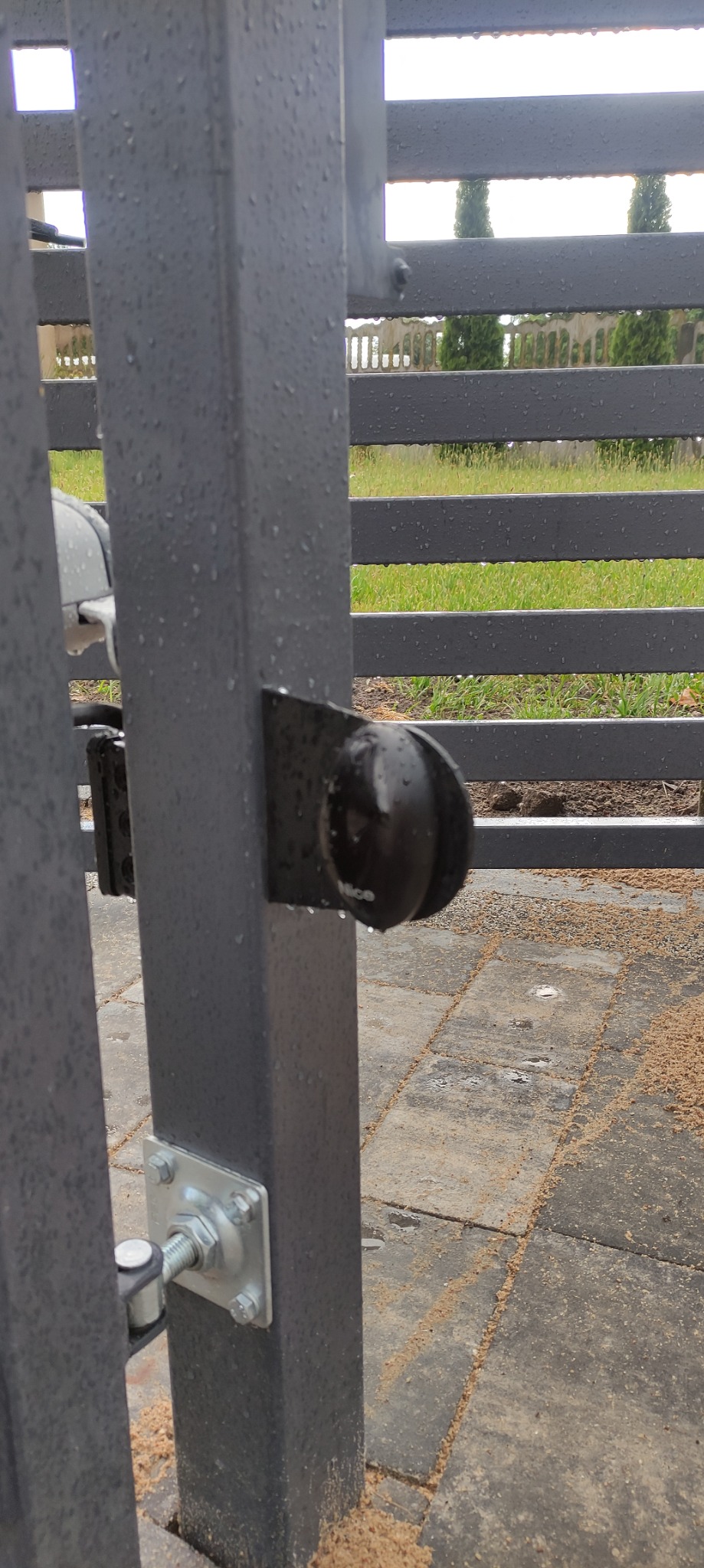 Szary słupek bramy z zamontowanym fotokomórką NICE, widoczne krople deszczu, w tle ogrodzenie panelowe i zieleń.