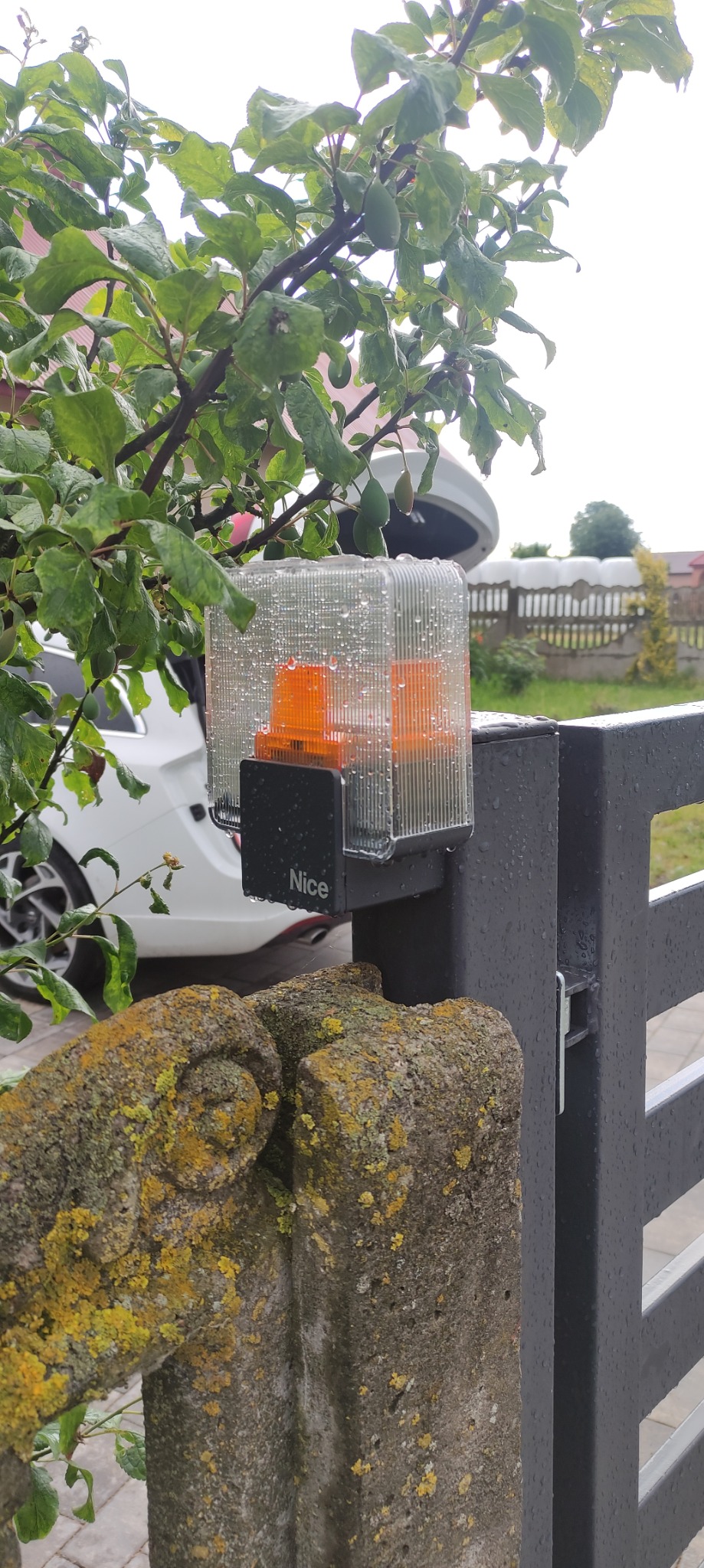 Lampa ostrzegawcza automatyki bramy Nice zamontowana na metalowym słupku bramy, widoczne krople deszczu na obudowie, w tle zielone drzewo i biały samochód z otwartą klapą bagażnika.