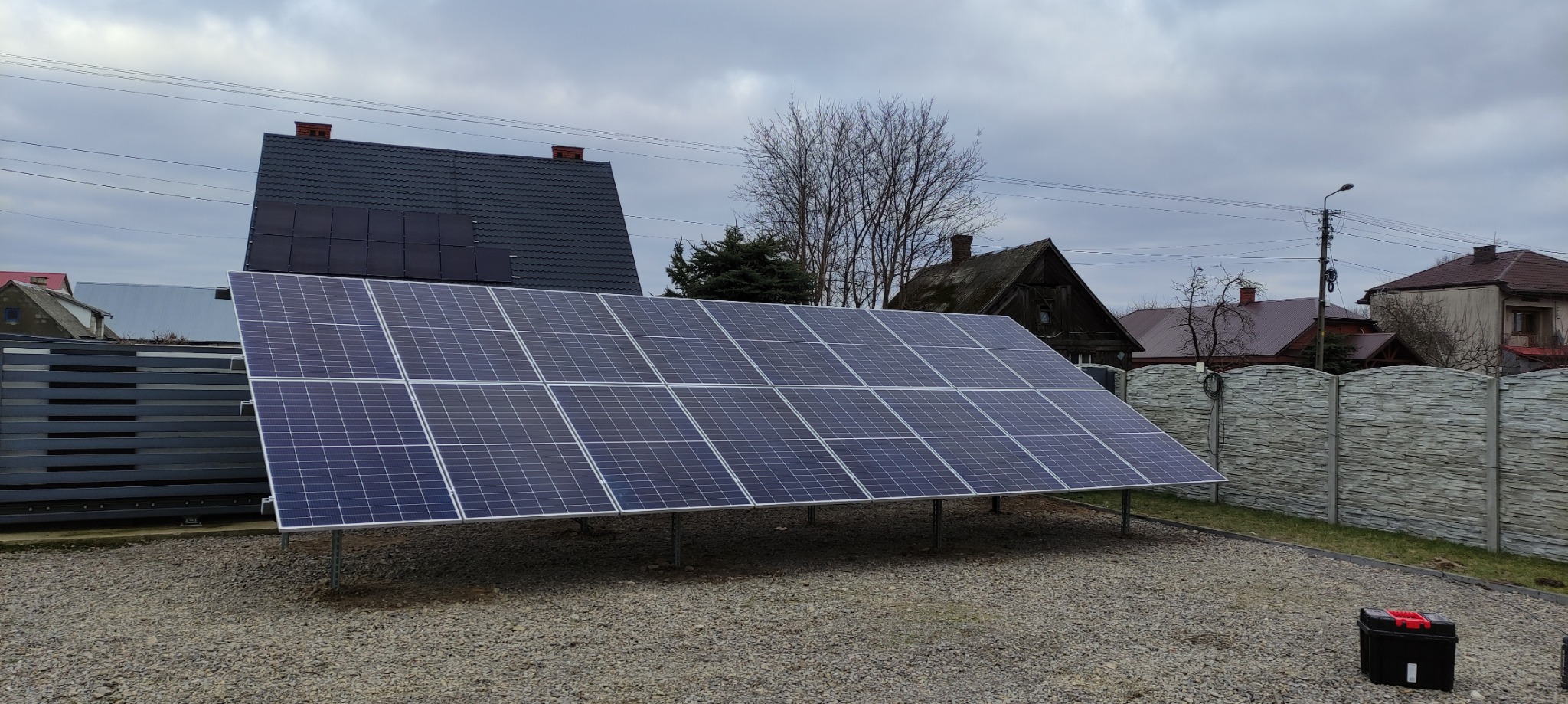 Panele fotowoltaiczne ustawione na gruncie przed domem, widoczny czarny kufer narzędziowy, pochmurne niebo w tle.