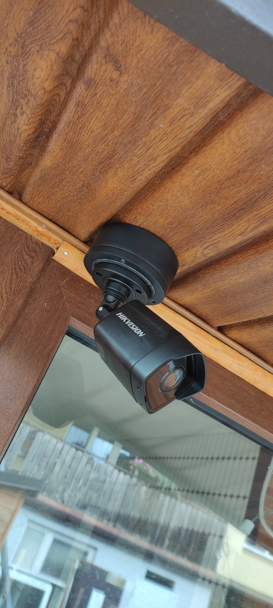Czarna kamera monitoringu Hikvision zamontowana pod drewnianym okapem, widoczna z boku, z odbiciem otoczenia w obiektywie.