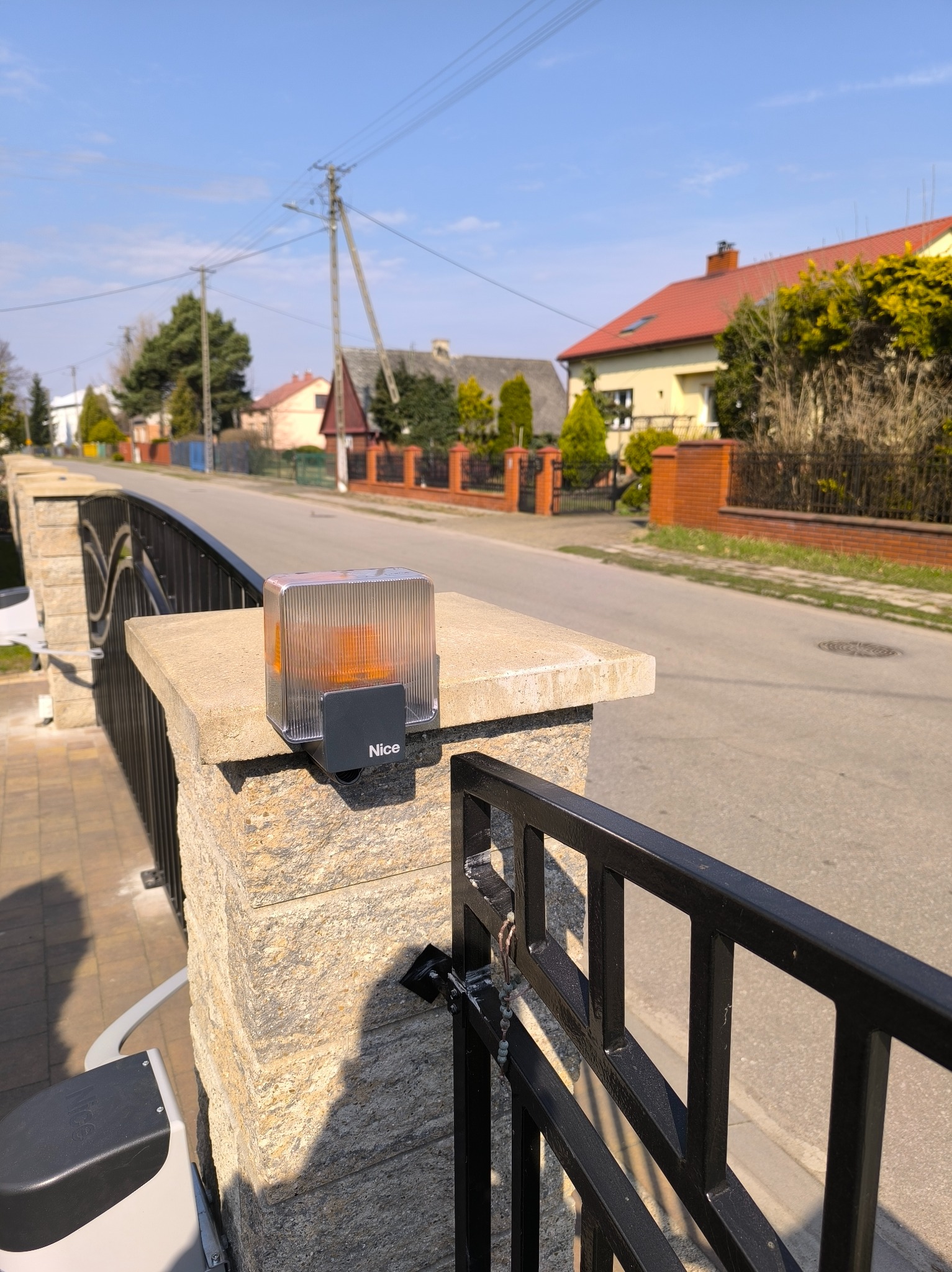 Lampa ostrzegawcza Nice zamontowana na słupku bramy wjazdowej, widok na ulicę i zabudowę jednorodzinną w tle. Słoneczny dzień.