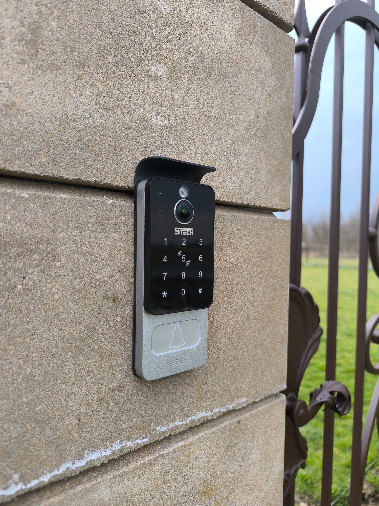 Zamontowany wideodomofon STEECH z klawiaturą numeryczną na murze obok metalowej bramy wjazdowej. Widoczne zielone tło ogrodu.