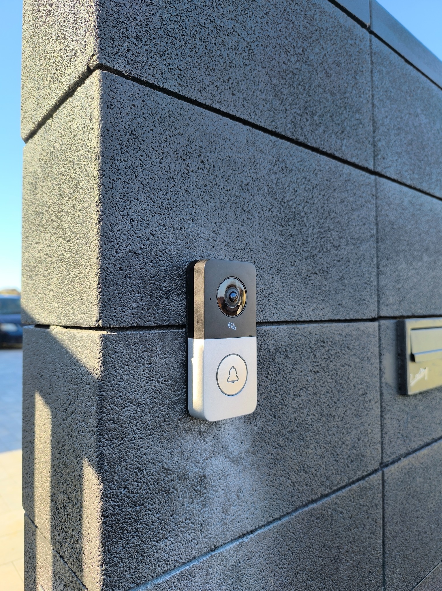 Nowoczesny wideodomofon z kamerą zamontowany na szarym, teksturowanym słupku ogrodzeniowym, z widoczną skrzynką na listy i fragmentem nieba w tle.