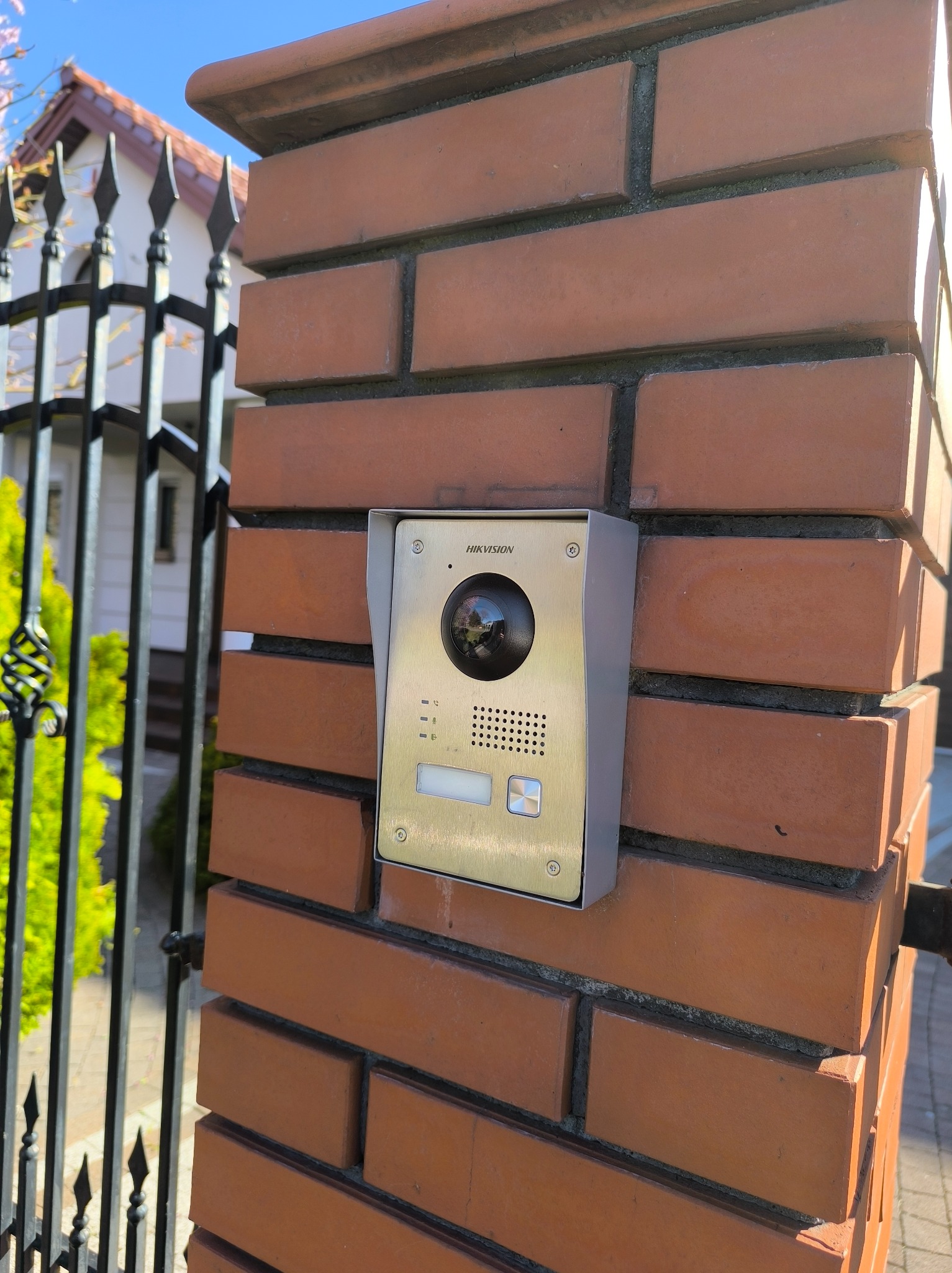 Złoto-srebrny wideodomofon Hikvision zamontowany na ceglanym słupku ogrodzenia, widok z boku, brama w tle, słoneczny dzień.